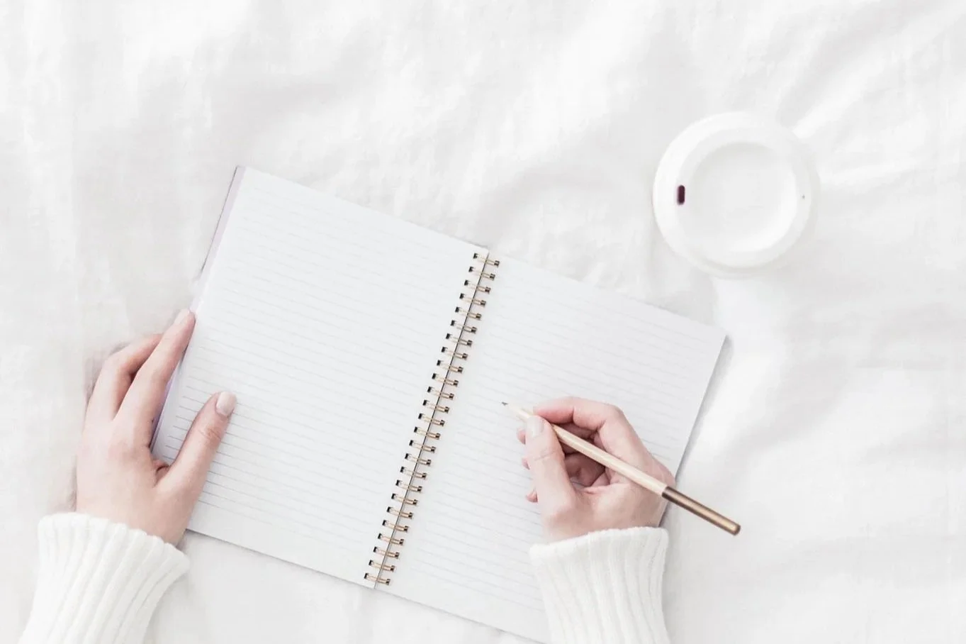 Minimal flat lay of hands writing in an open notebook on white bedding — quiet workspace, creative planning for business launch.