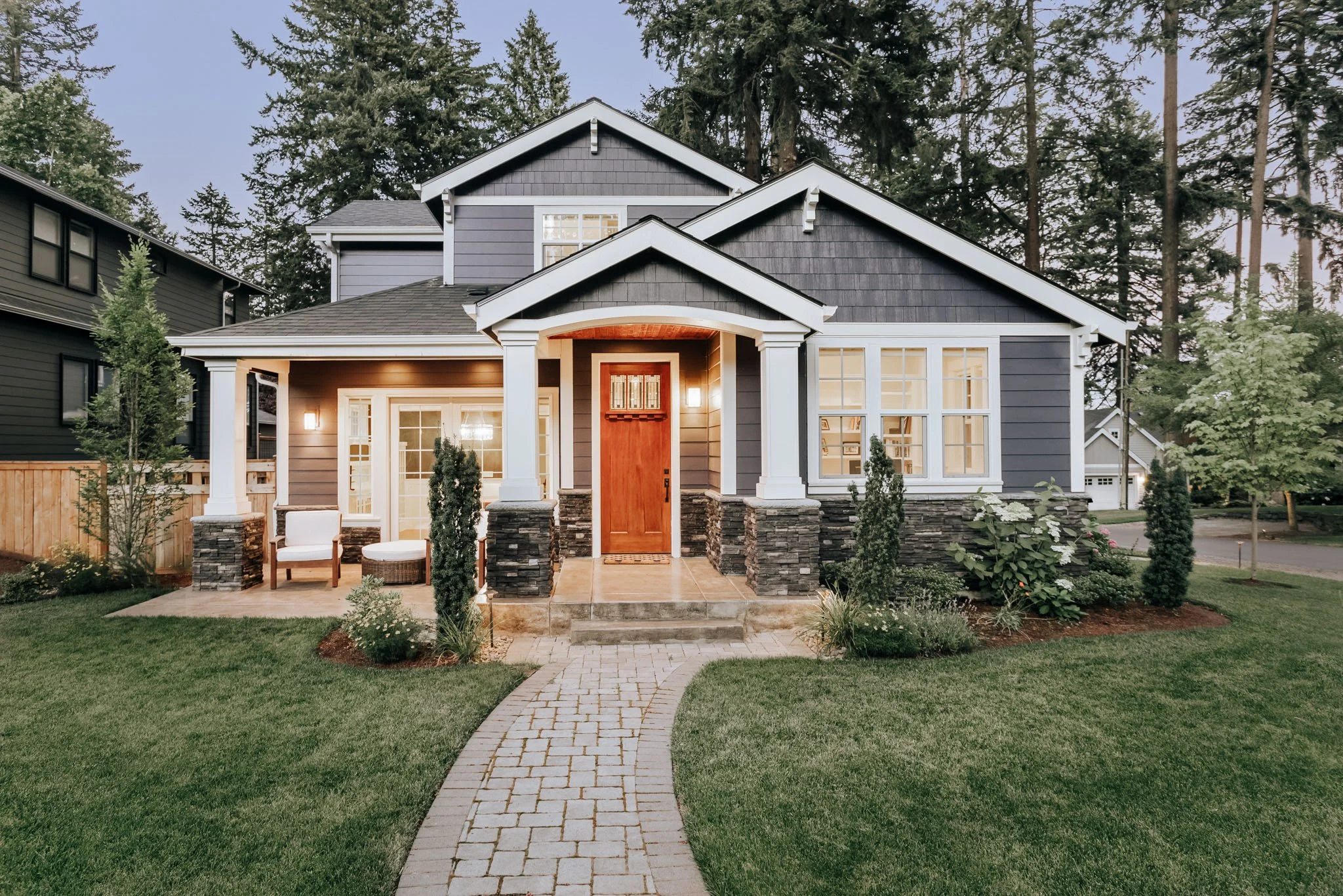 “A classic American Craftsman-style home with a deep front porch, tapered columns, shingle siding, warm exterior lighting, and a landscaped garden path leading to a wooden front door.”