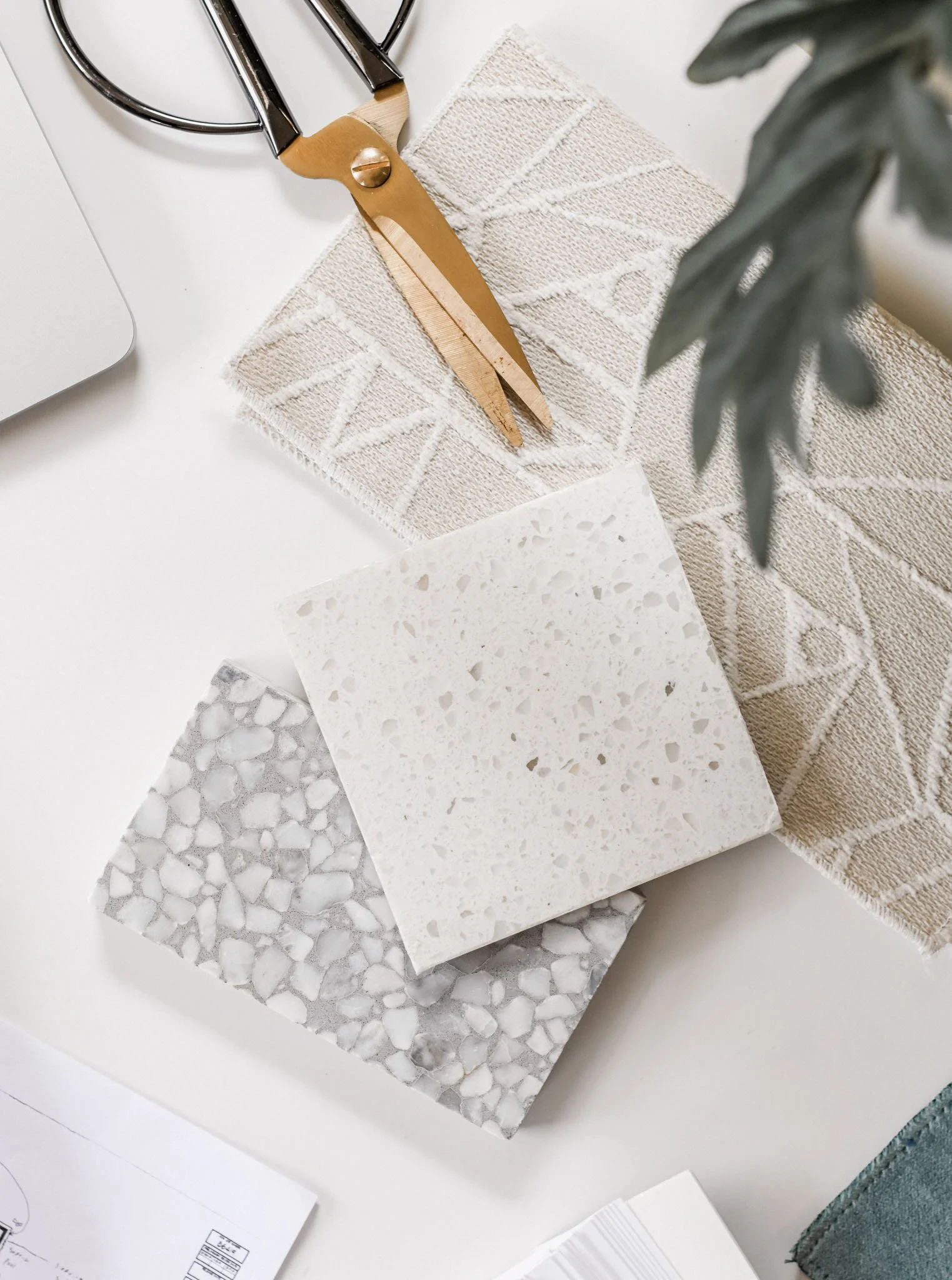 Neutral terrazzo and cream stone samples laid beside textured upholstery swatches and brass scissors — a clean, design-led renovation palette arranged on a white desk.