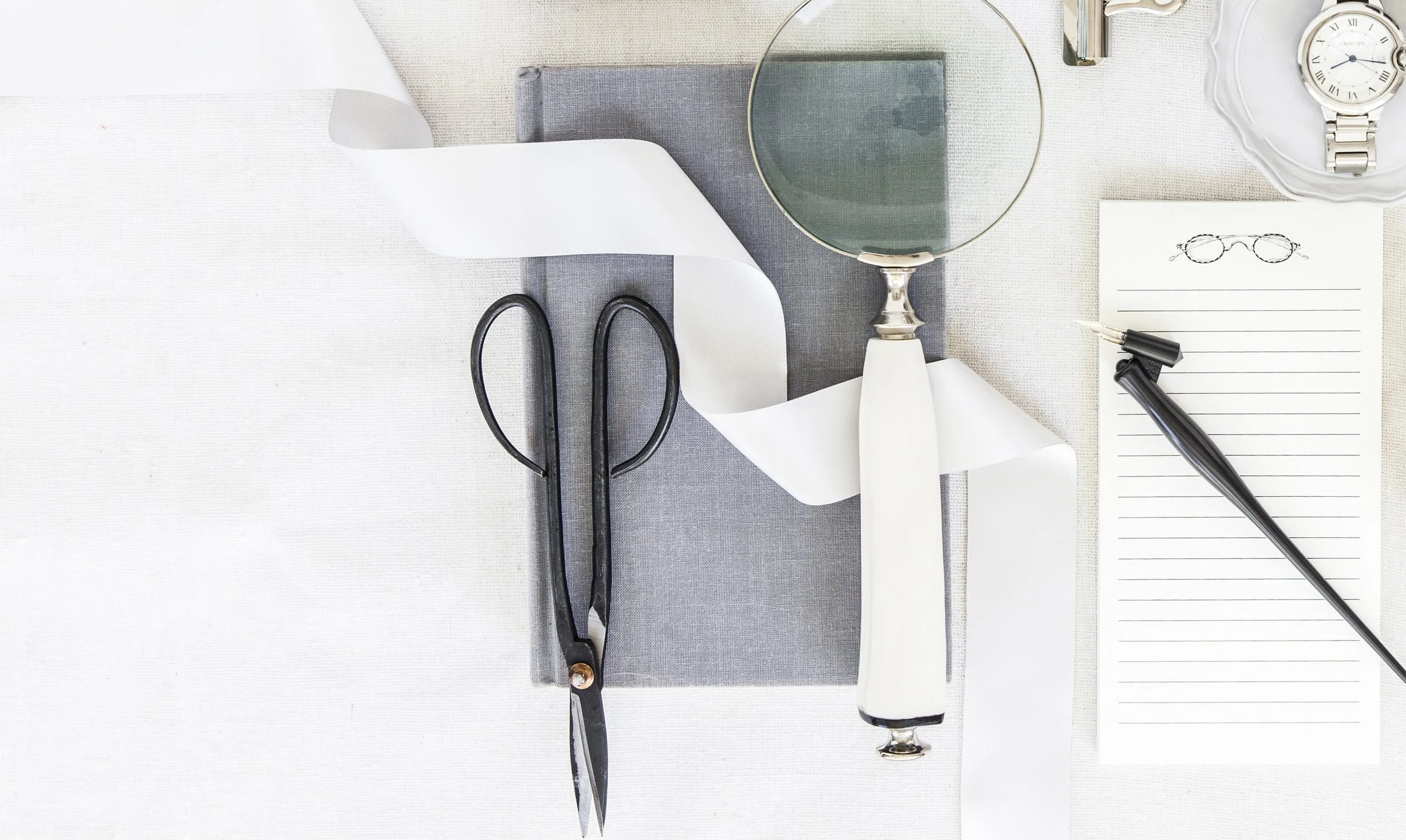 Neutral flatlay of design tools — scissors, ribbon, magnifying glass and notebook arranged on linen — expressing the materiality and structure behind packaging design.