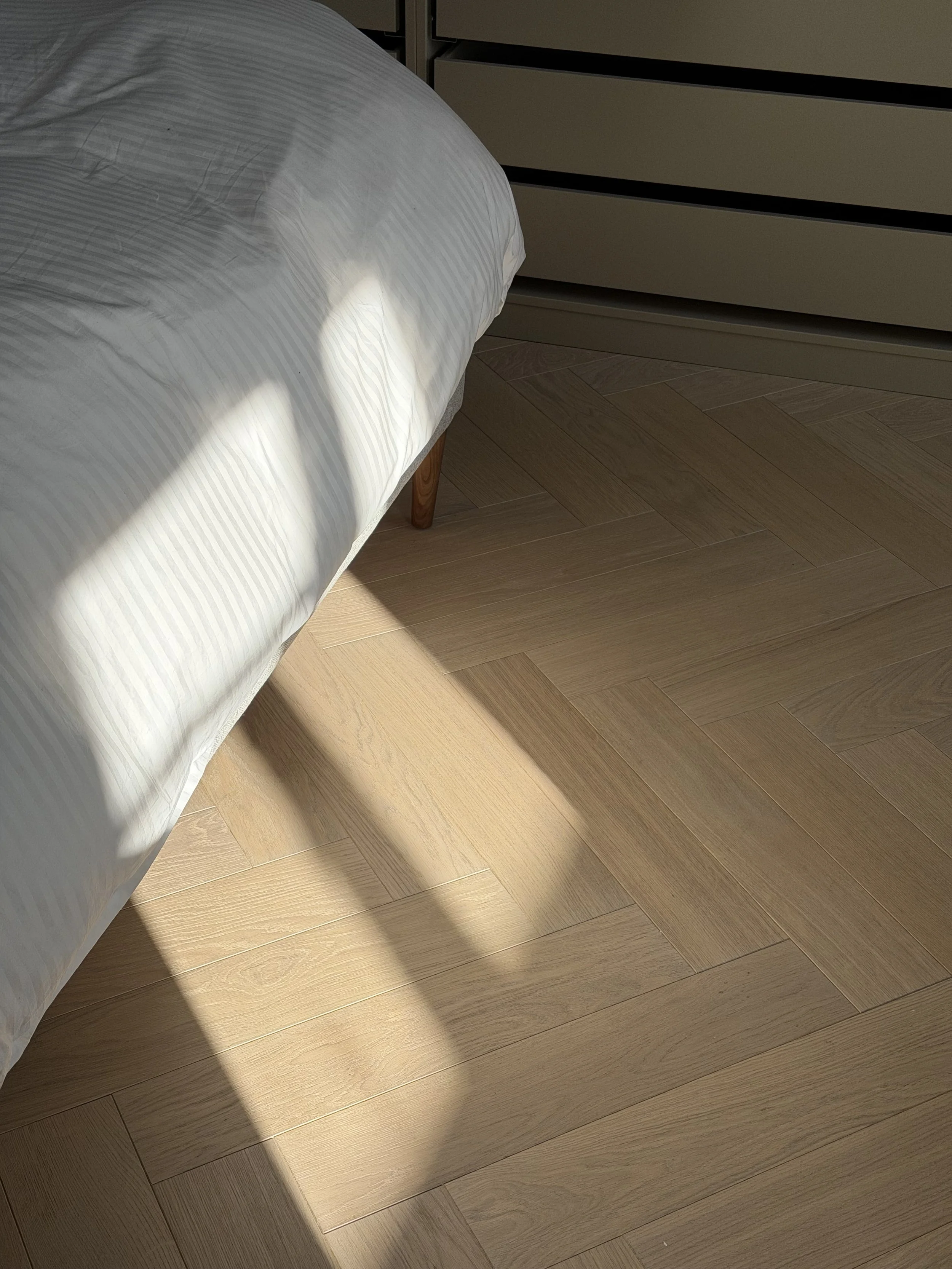 Soft morning light falling across a herringbone wood floor beside a white duvet — a quiet, atmospheric moment showing how light shifts between seasons and places.