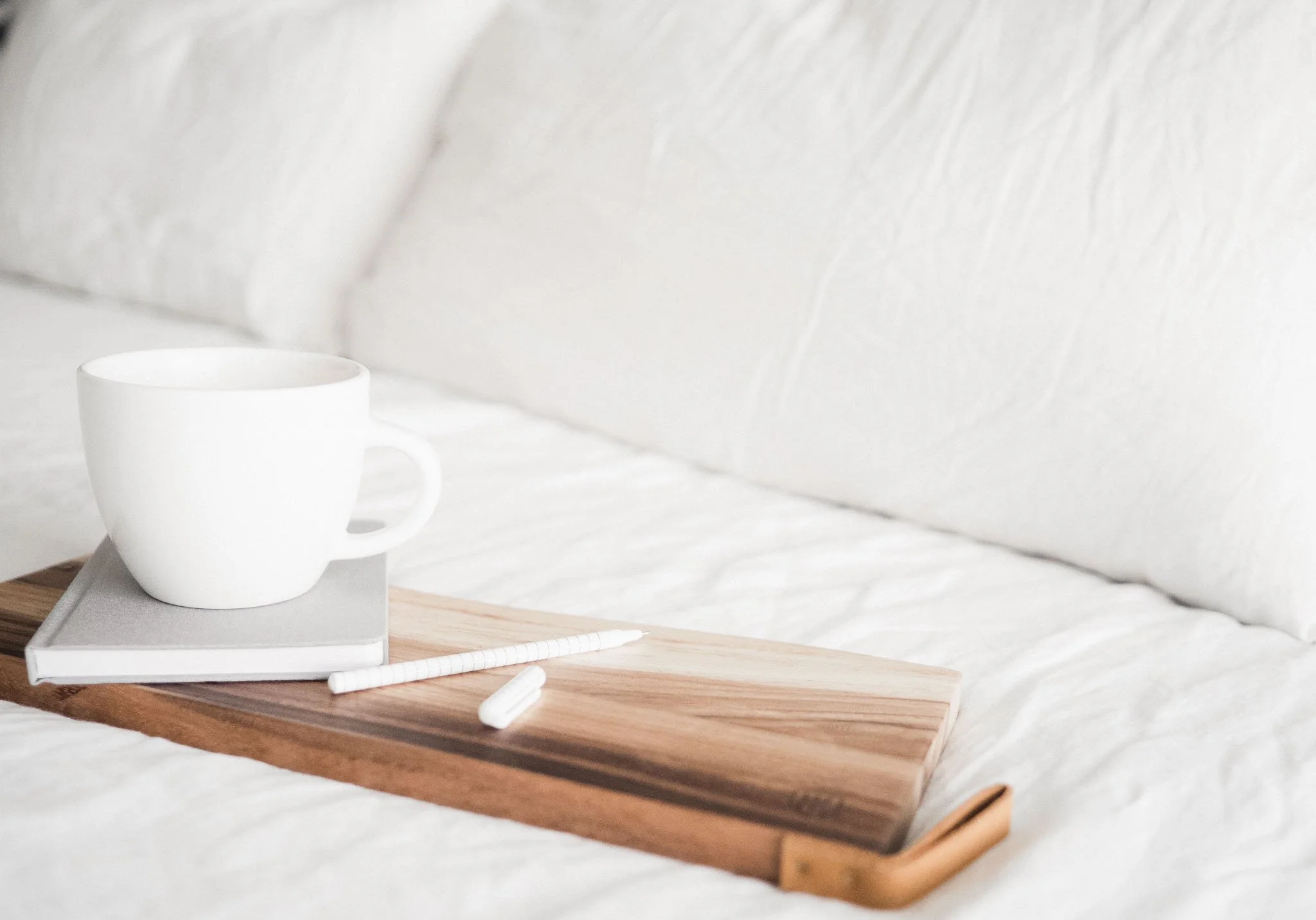 White coffee mug and notebook styled on a wooden tray over soft white bedding — a quiet, functional morning scene in a calm, minimal home interior.