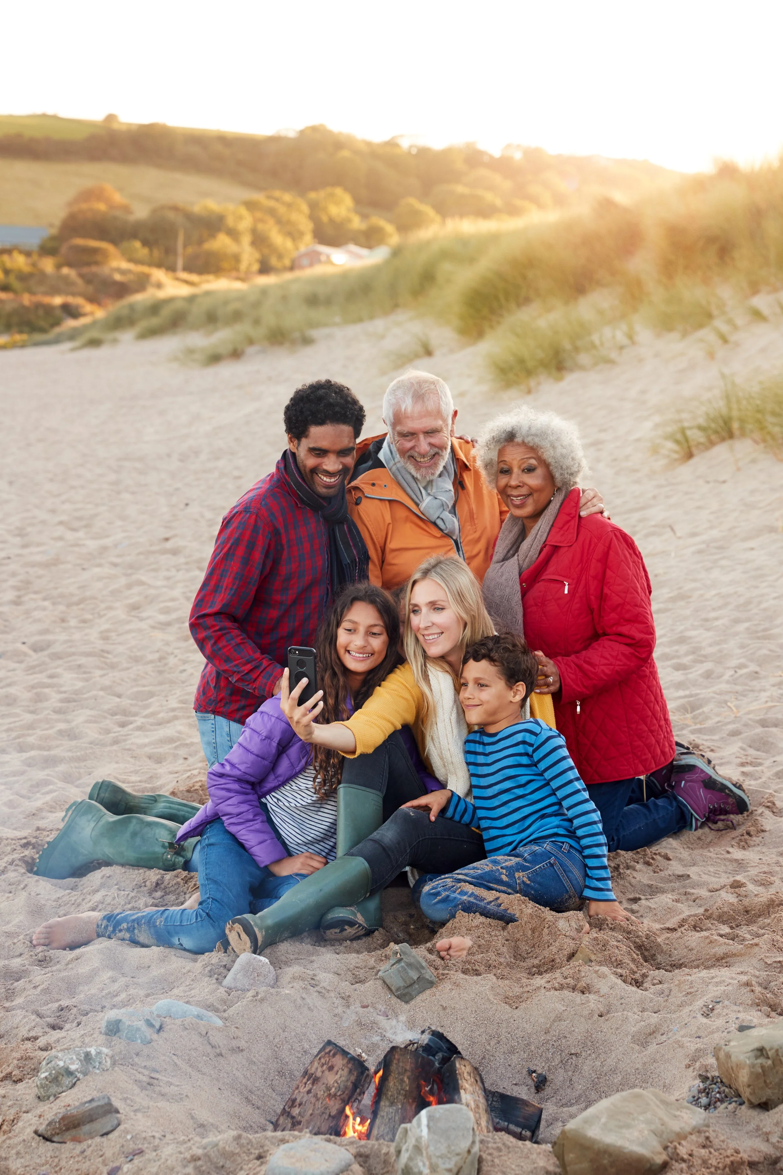 Multigenerational family sitting together on a beach around a campfire, smiling and taking a selfie, representing connection and healing in family therapy.