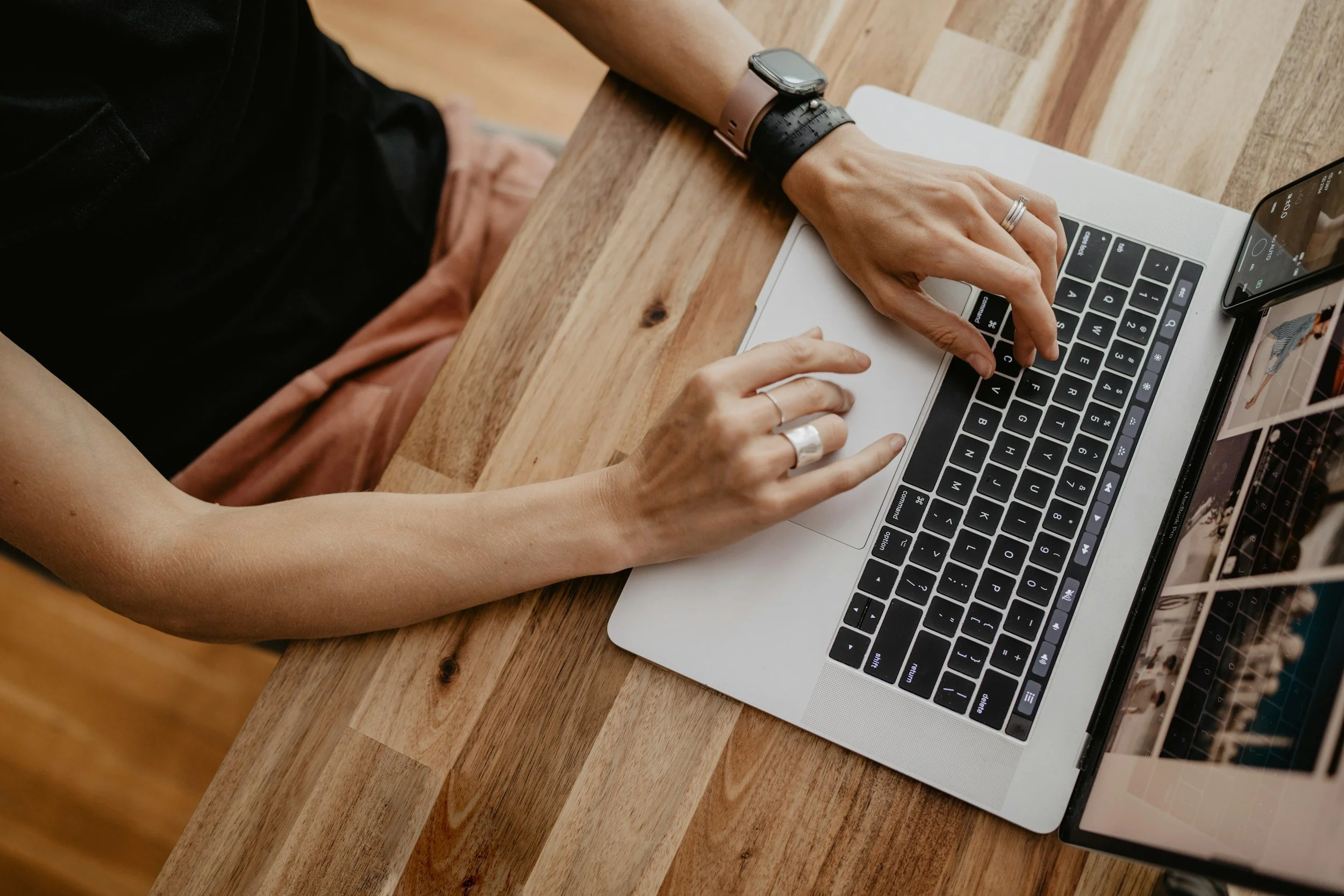 Person using a laptop on a wooden table, wearing a smartwatch and rings.