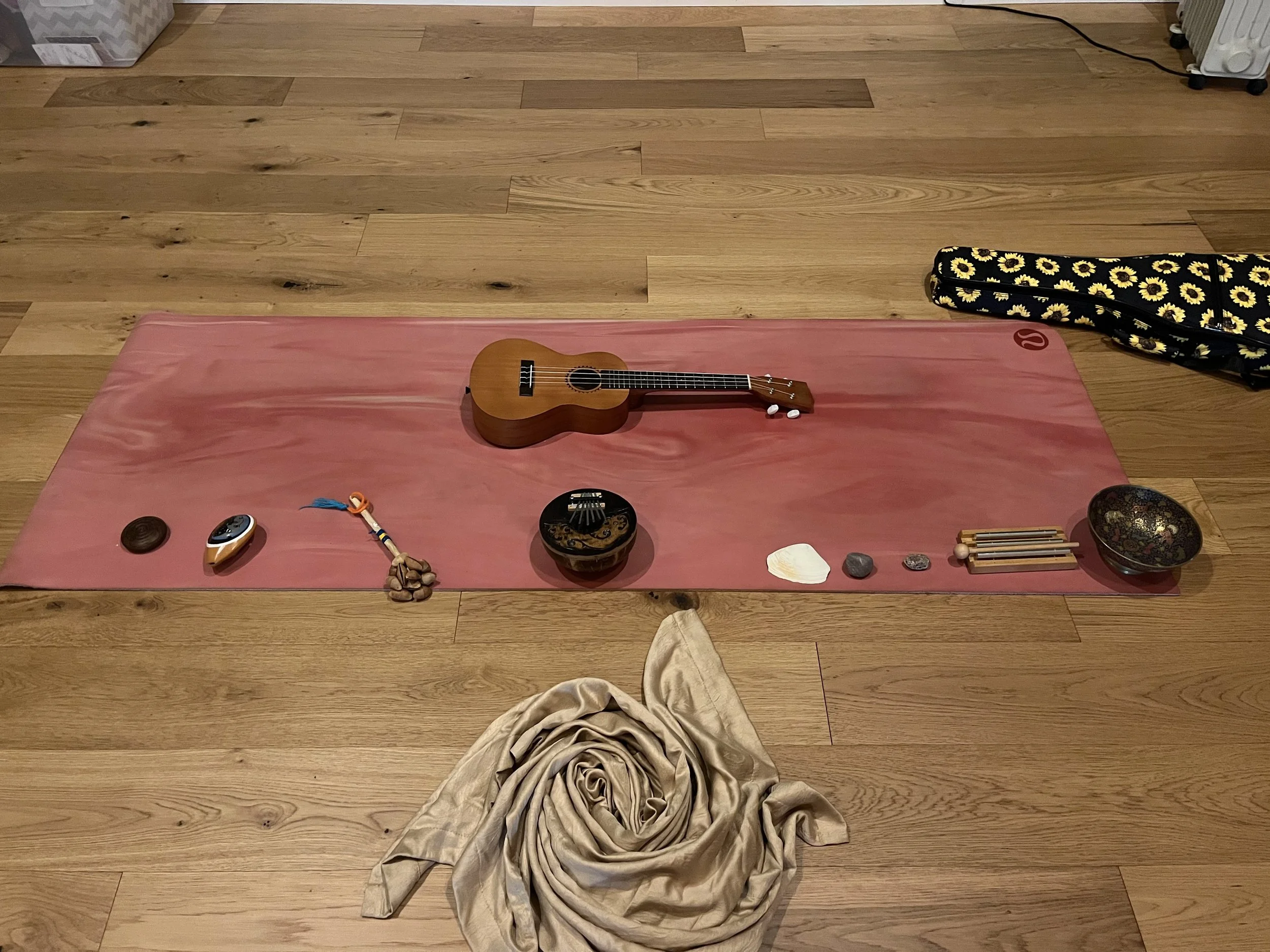 A yoga mat with musical instruments and objects for meditation, including a ukulele, a sound bowl, stones, and a cloth on a wooden floor.
