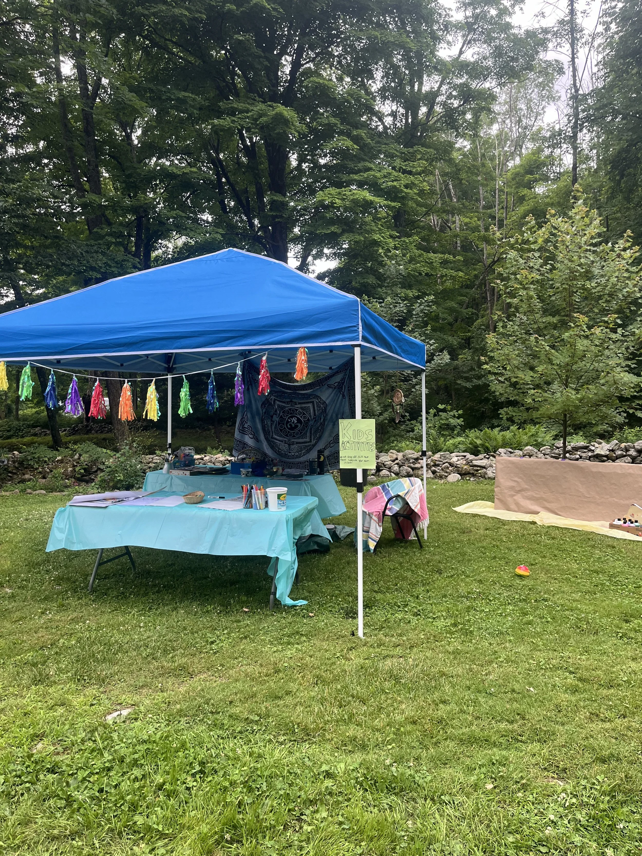 Outdoor event setup with a blue canopy and a table covered with a light blue cloth. The table has art supplies, papers, and a sign indicating 'Kids Activities.' Colorful tassels decorate the canopy, and there's a natural backdrop of green trees and g