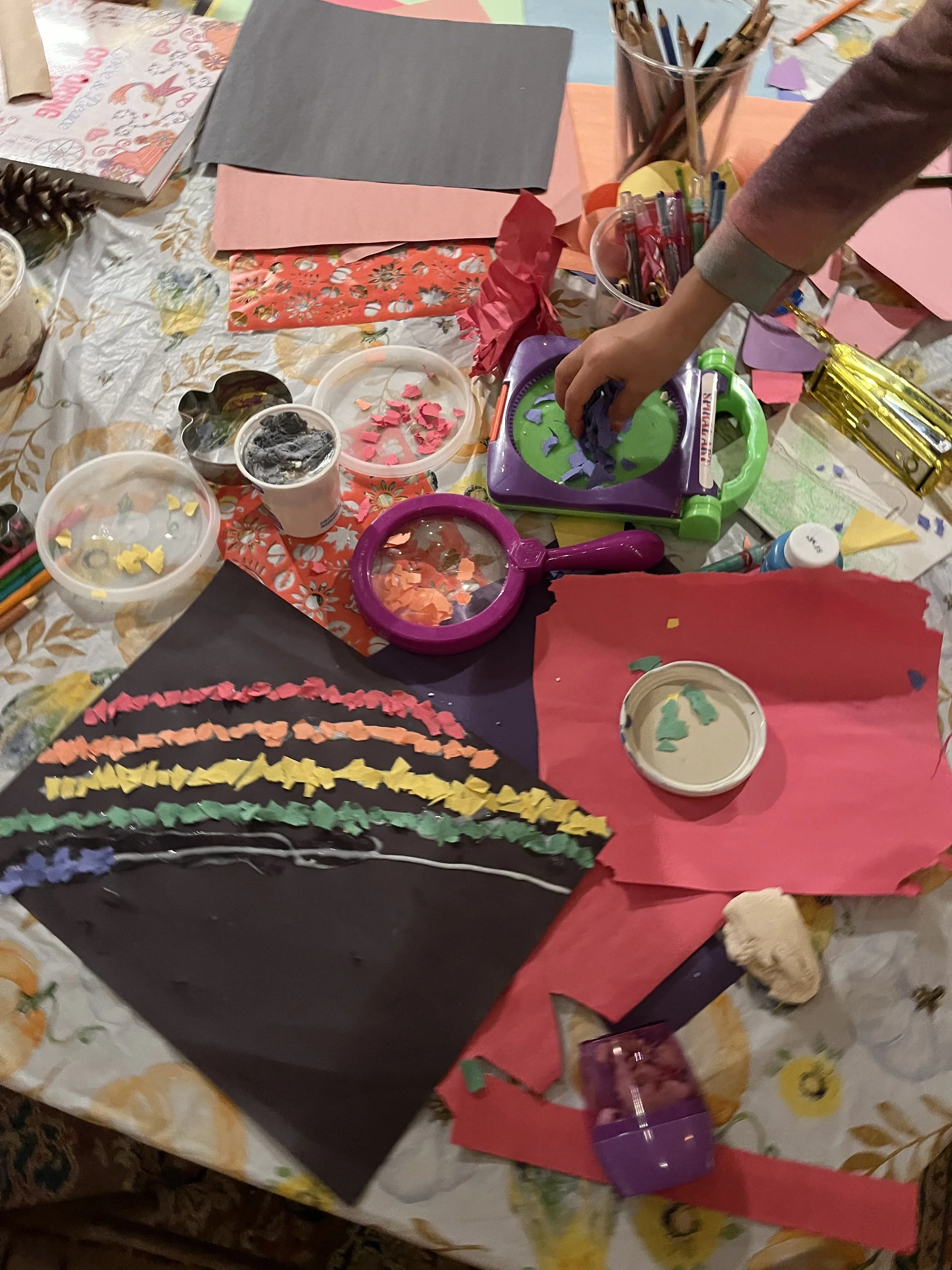 Art and craft supplies on a table, including colored paper, scissors, paper shreds, and drawing tools. A hand is holding some paper pieces.