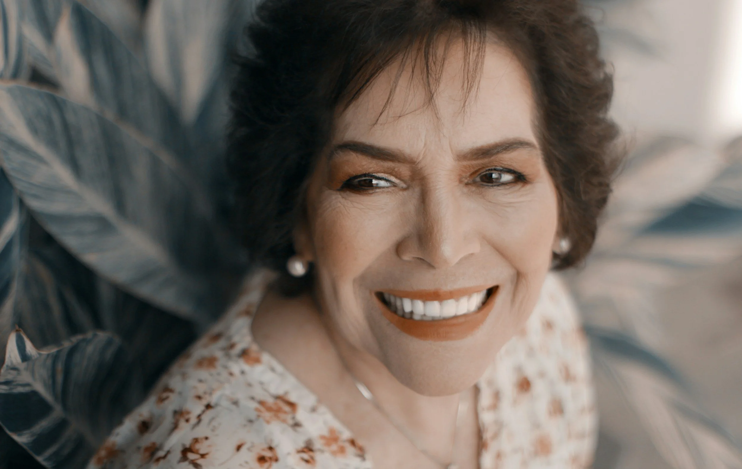 Close-up of a smiling woman with short dark hair, wearing pearl earrings and a floral blouse, surrounded by large tropical leaves.