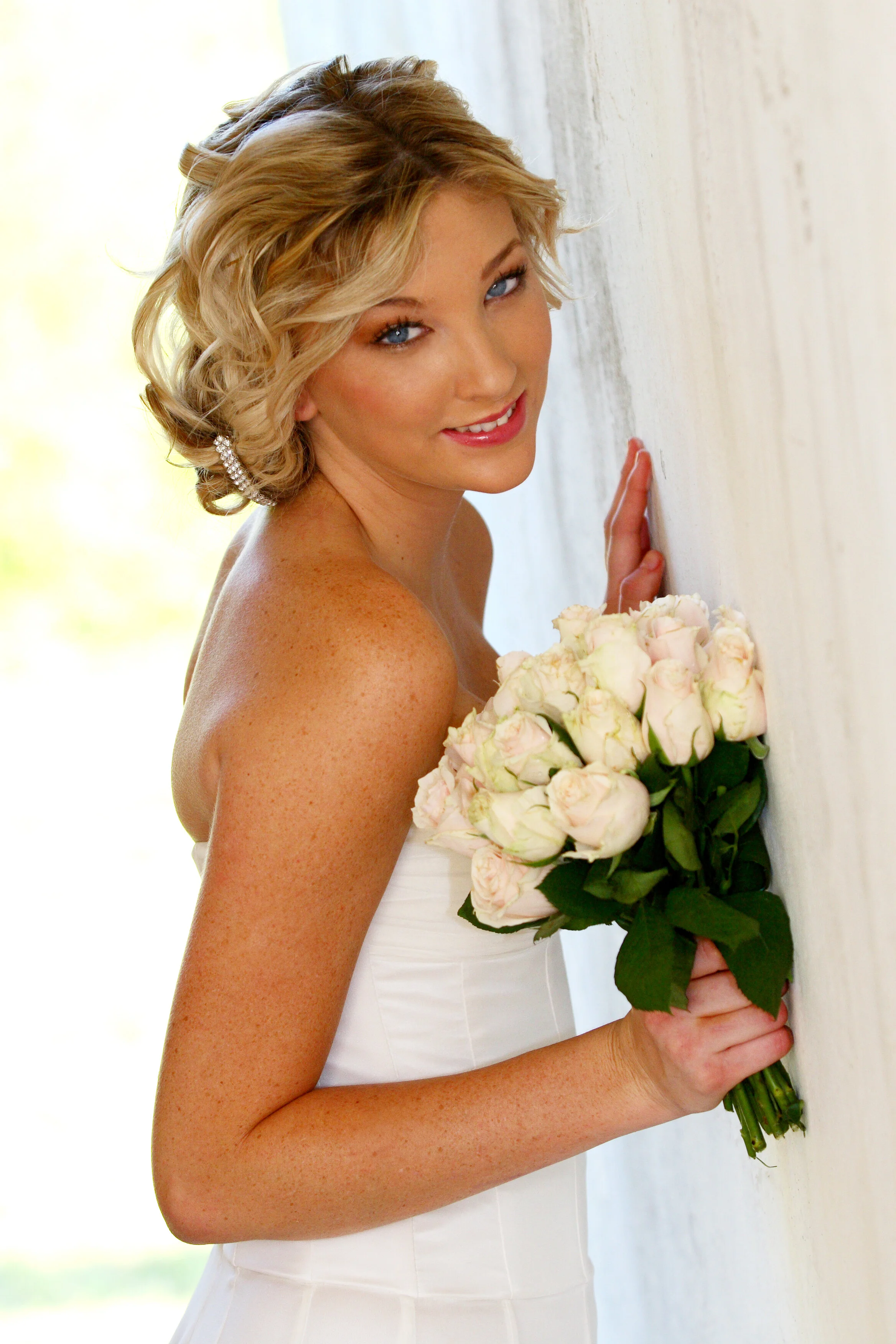A blonde woman in a strapless white dress holding a bouquet of pale pink roses, smiling and leaning against a light-colored wall.