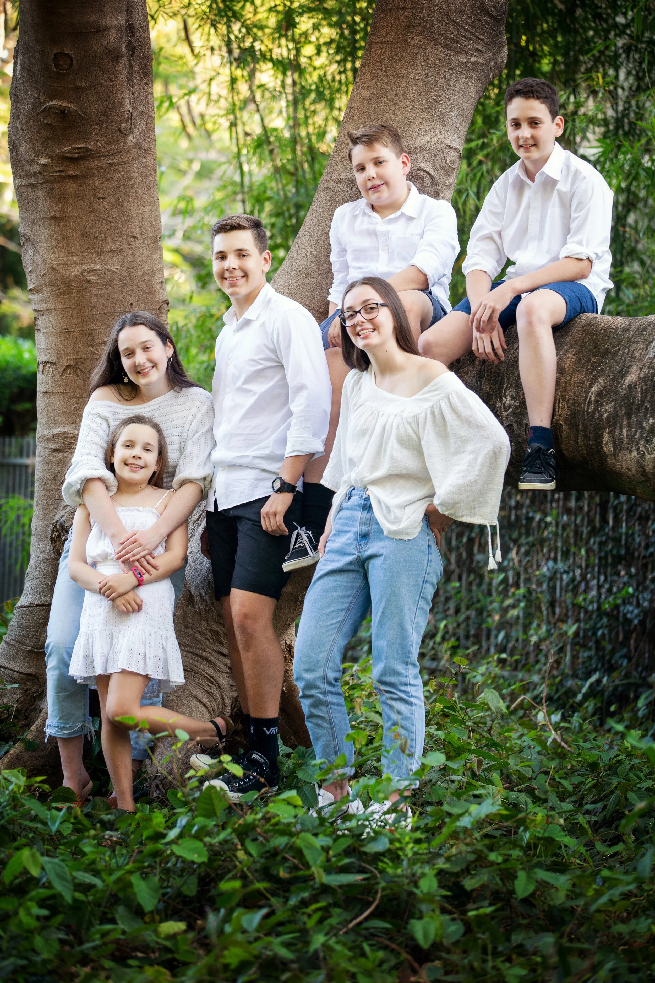 A group of seven children, three girls and four boys, gathered outdoors around a large tree in a lush green forest setting, smiling at the camera.