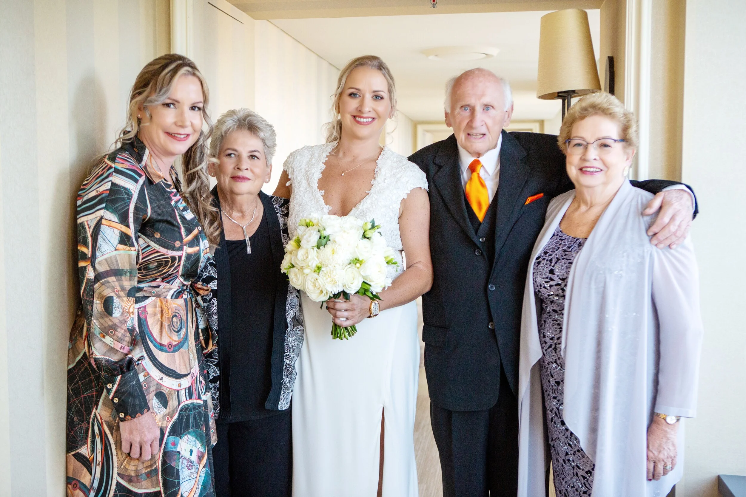 Group of six women, including a bride holding a bouquet, standing together inside a room with beige walls and a floor lamp in the background.
