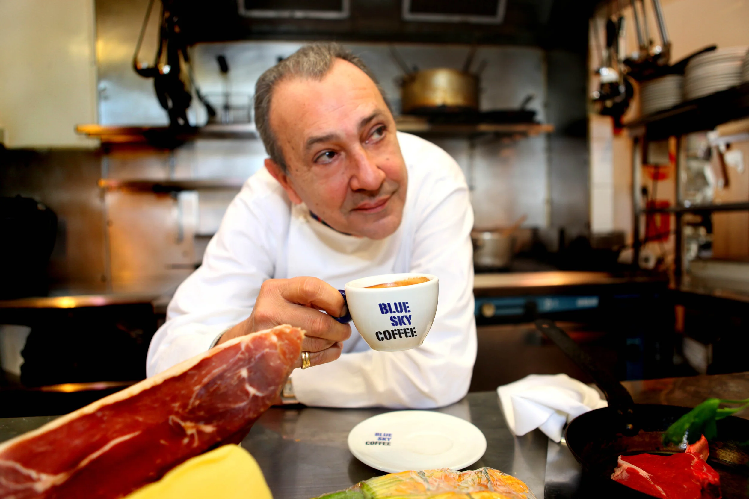 A chef in a white uniform holding a blue and white coffee cup labeled 'Blue Sky Coffee' in a professional kitchen.