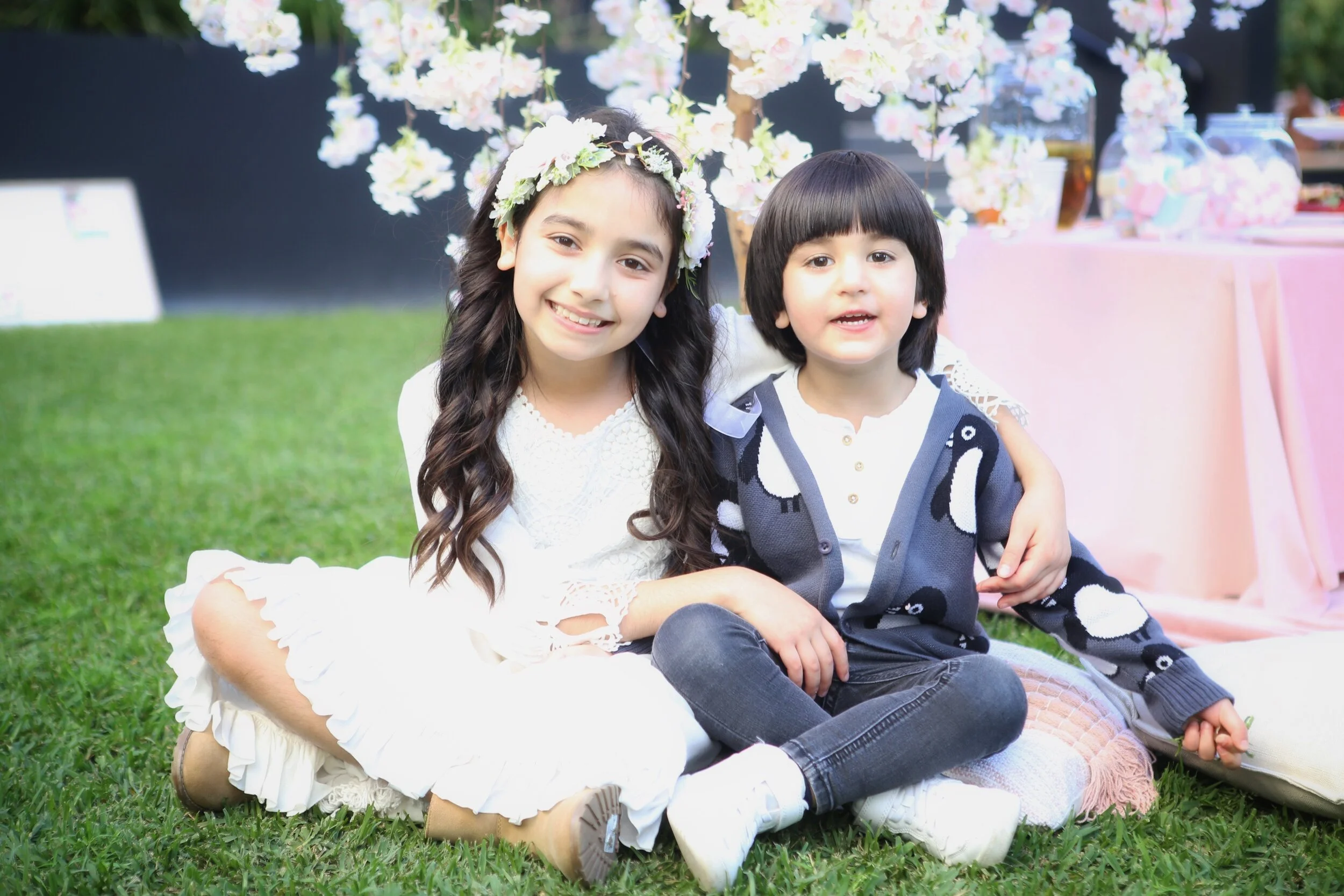 Two children sitting on grass at a party, one girl with long dark hair wearing a flower crown and a white dress, and one boy with short dark hair wearing a penguin patterned cardigan, both smiling.