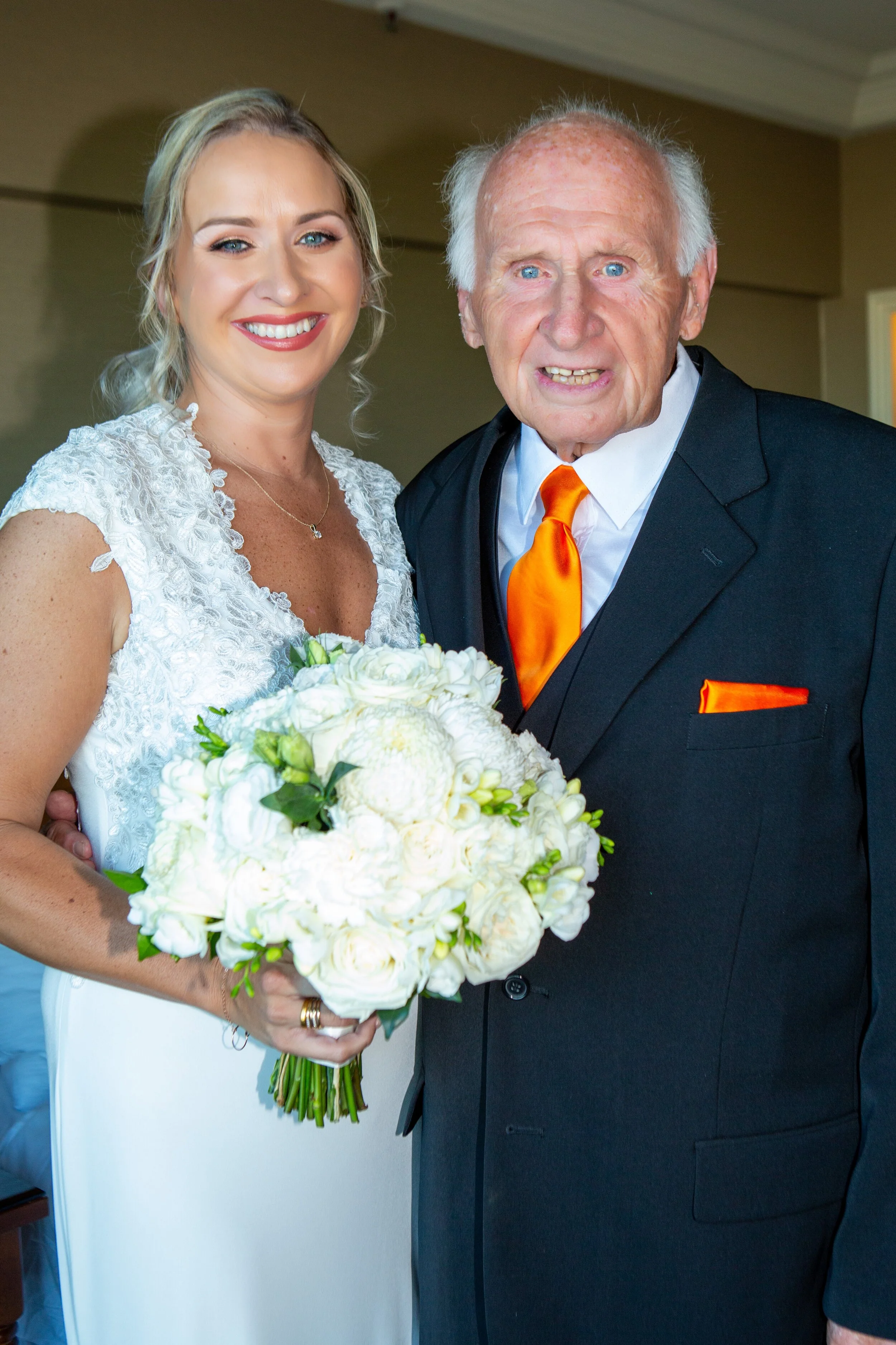 A young woman in a white wedding dress holding a bouquet of white flowers, standing next to an elderly man dressed in a black suit with an orange tie and pocket square, inside a room.