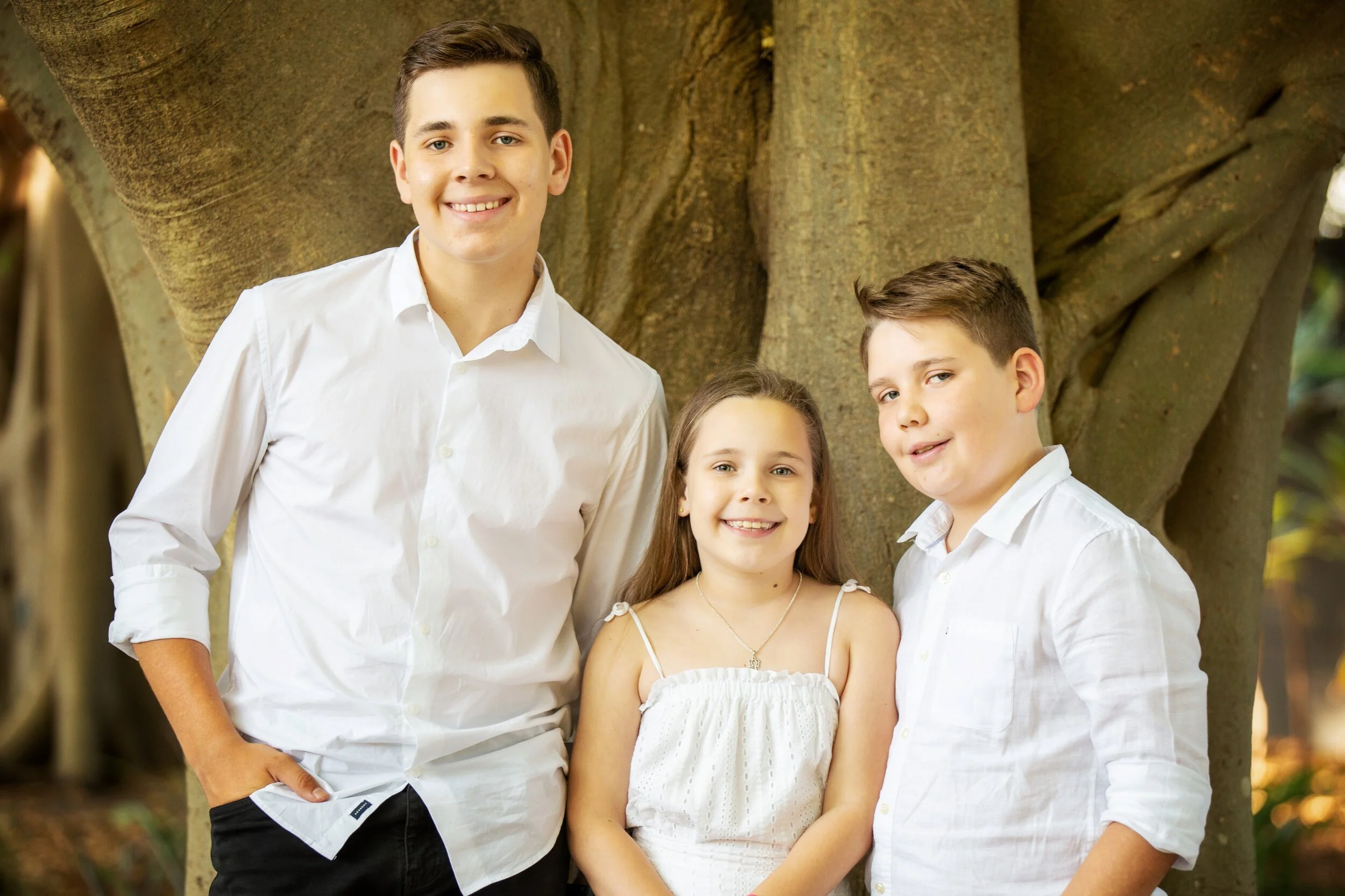 Three children, two boys and one girl, smiling outdoors in front of a large tree, dressed in white shirts and a white dress.