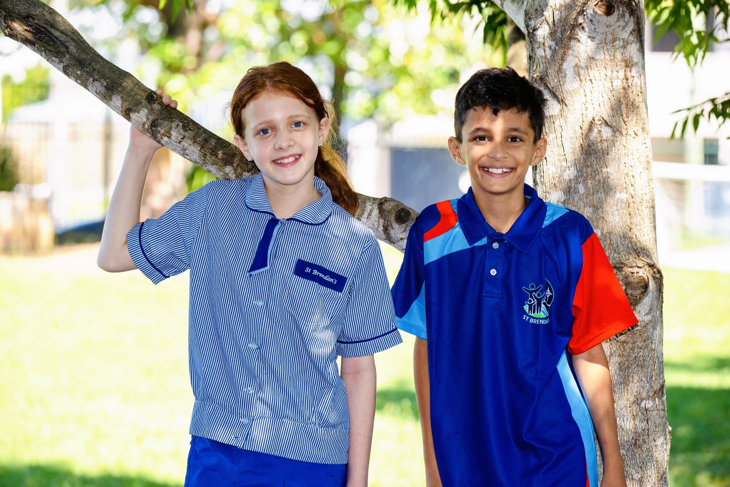 Two children, a girl with red hair and a boy with dark hair, smiling and standing outdoors beside a tree. The girl is wearing a blue school uniform and the boy a blue and red sports uniform.