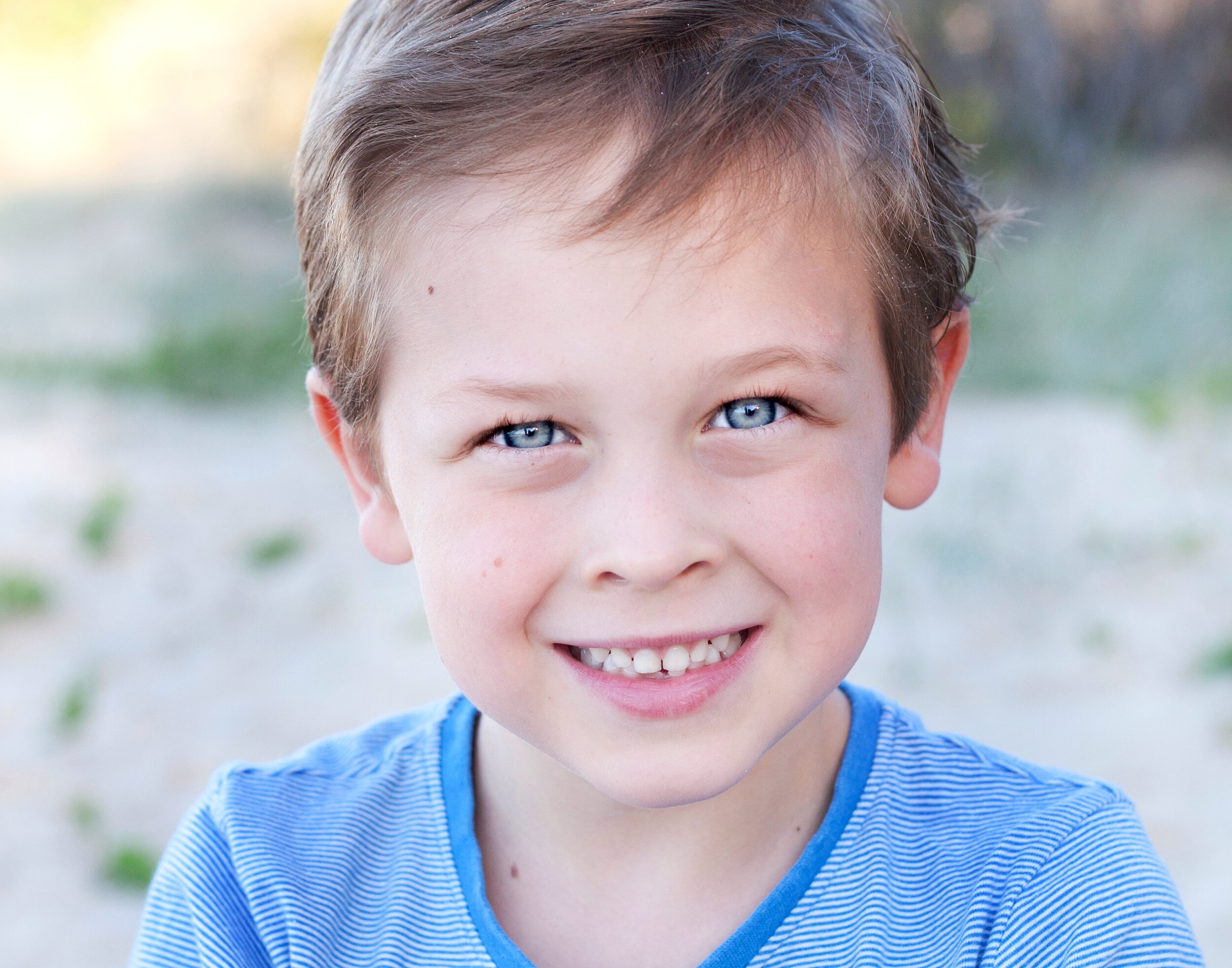Close-up of a smiling young boy with blue eyes and short brown hair, wearing a blue striped shirt outdoors.
