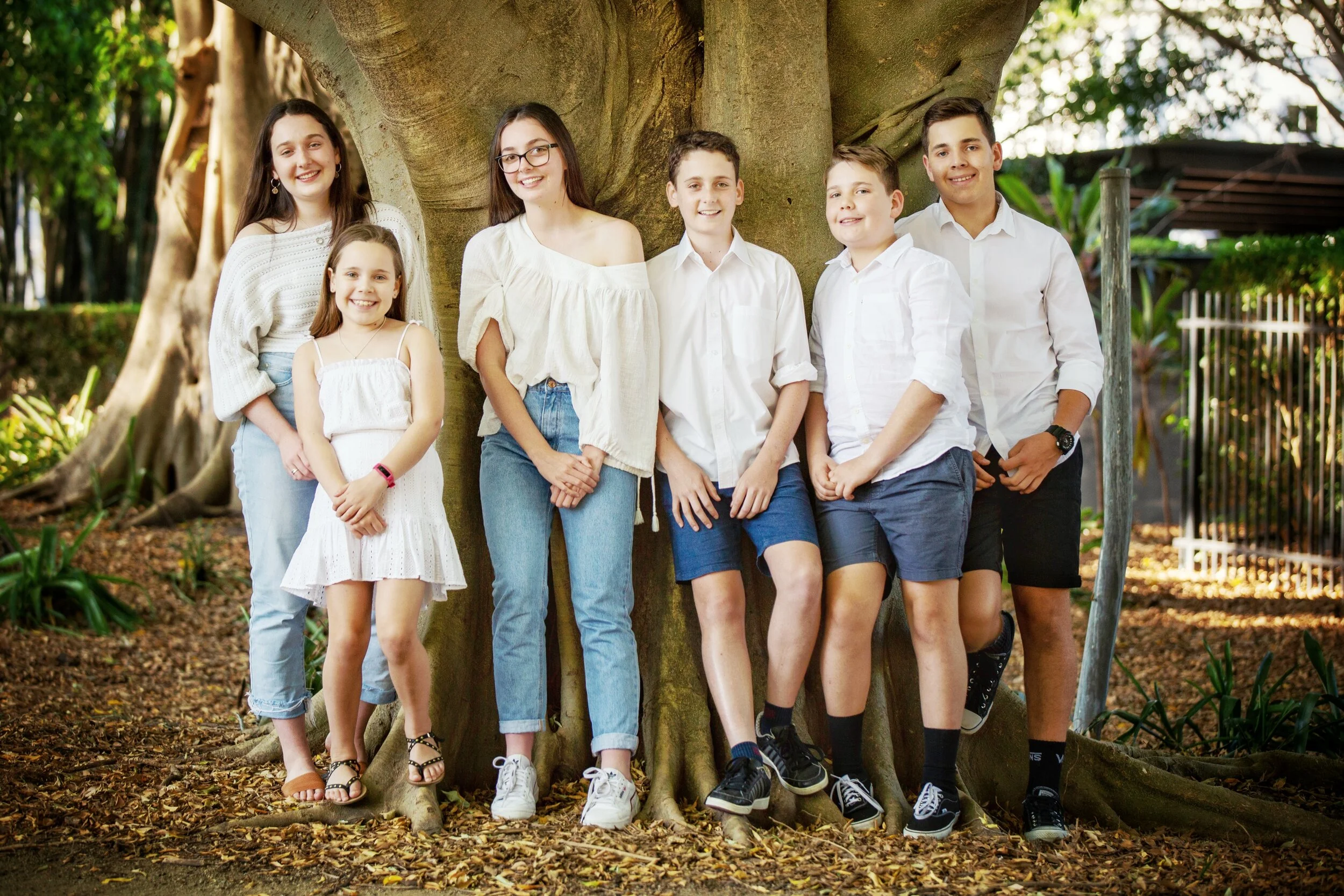 Group of seven children standing in front of a large tree with green foliage in the background.
