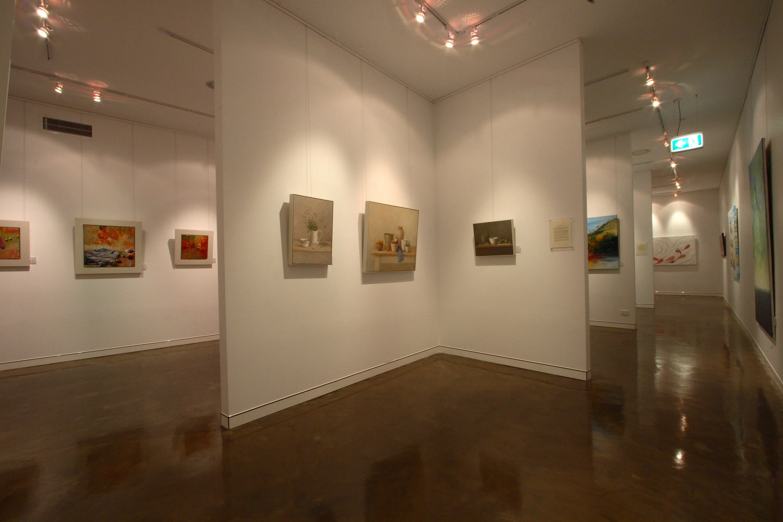 Art gallery with white walls displaying various paintings and artworks, polished dark wooden floor, and ceiling track lighting.