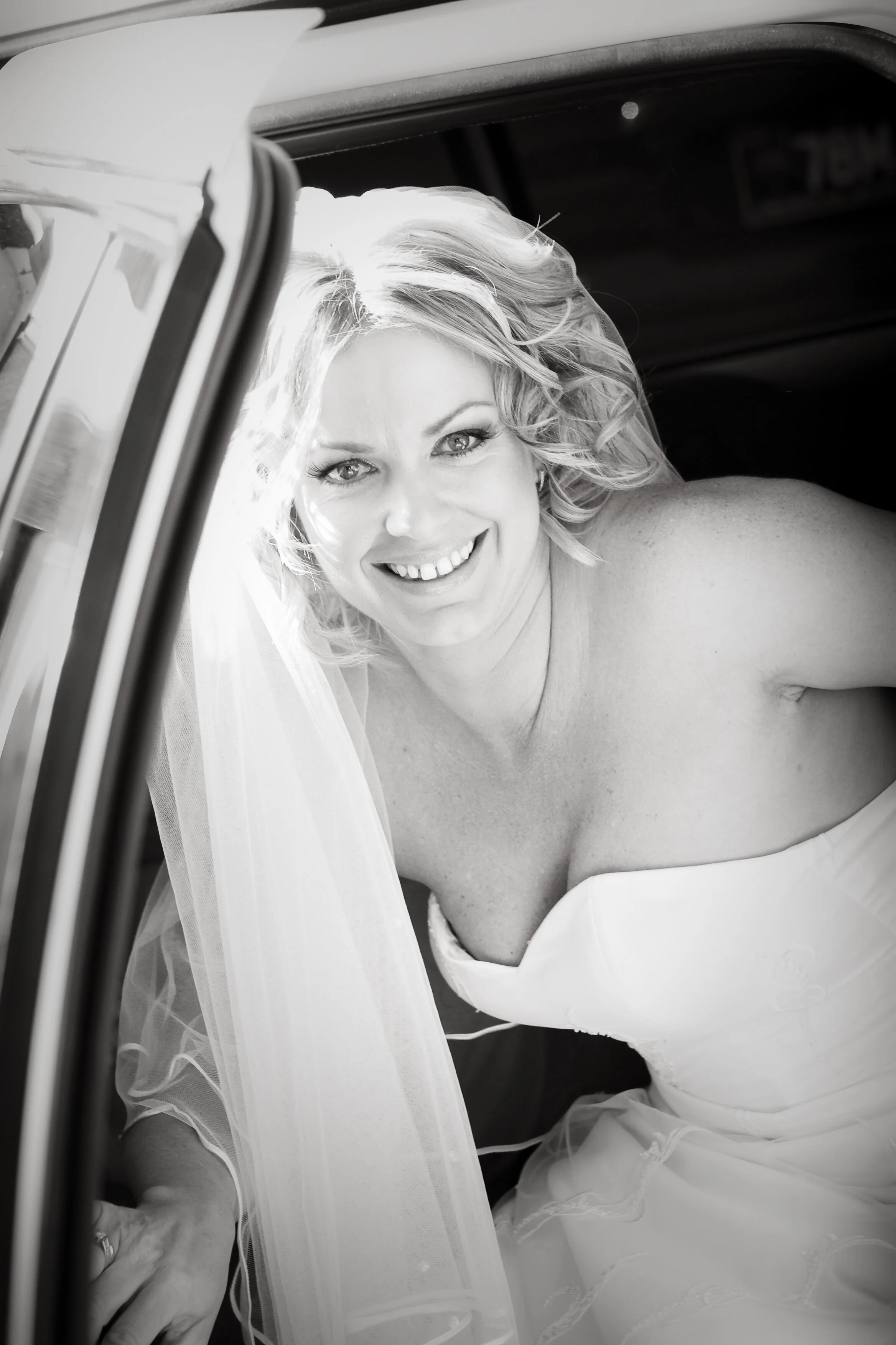 Black and white photo of a smiling bride with wavy blonde hair, wearing a strapless wedding dress and veil, leaning into a car with the door open.