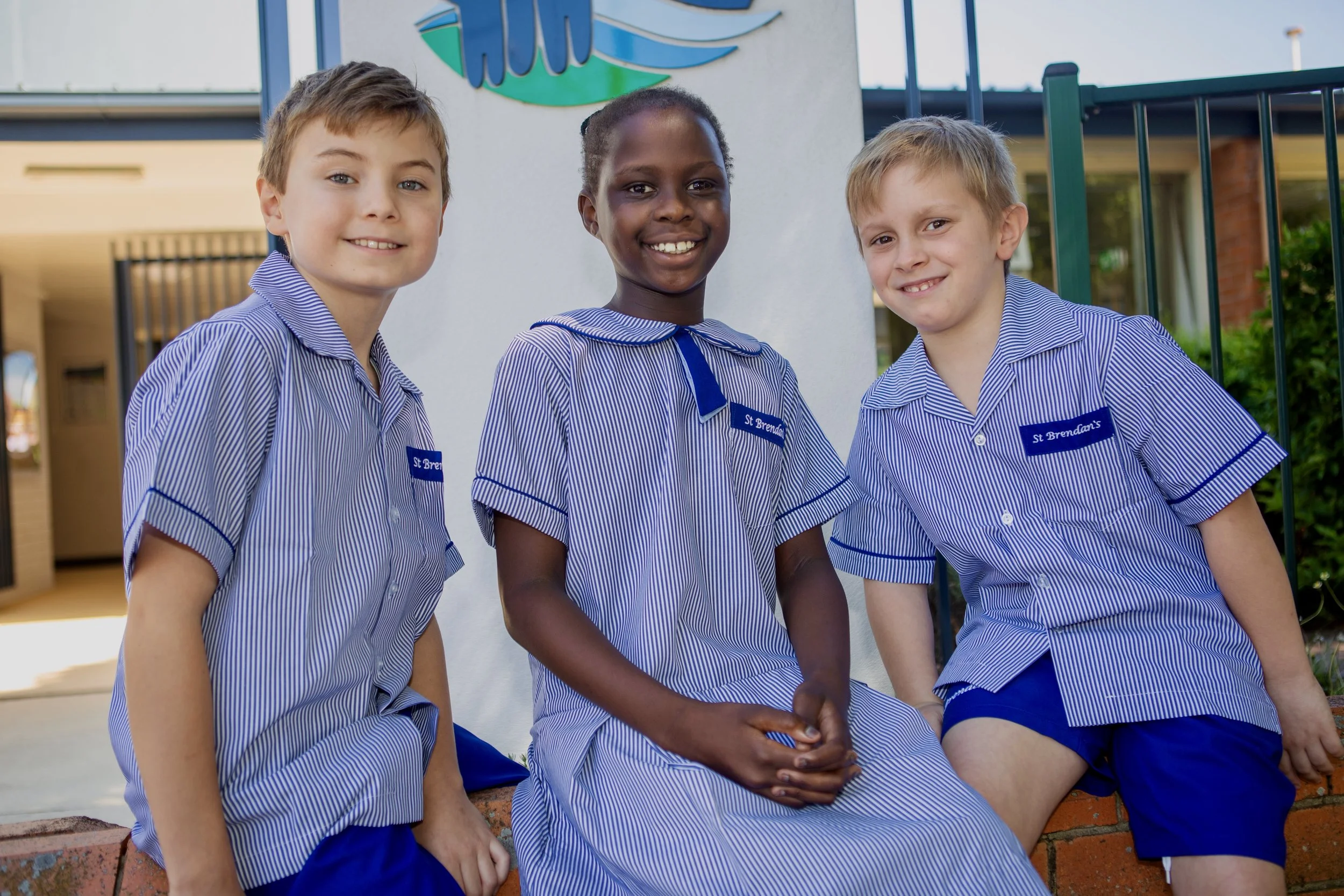 Three children in school uniforms sitting outside in front of a building, smiling at the camera.