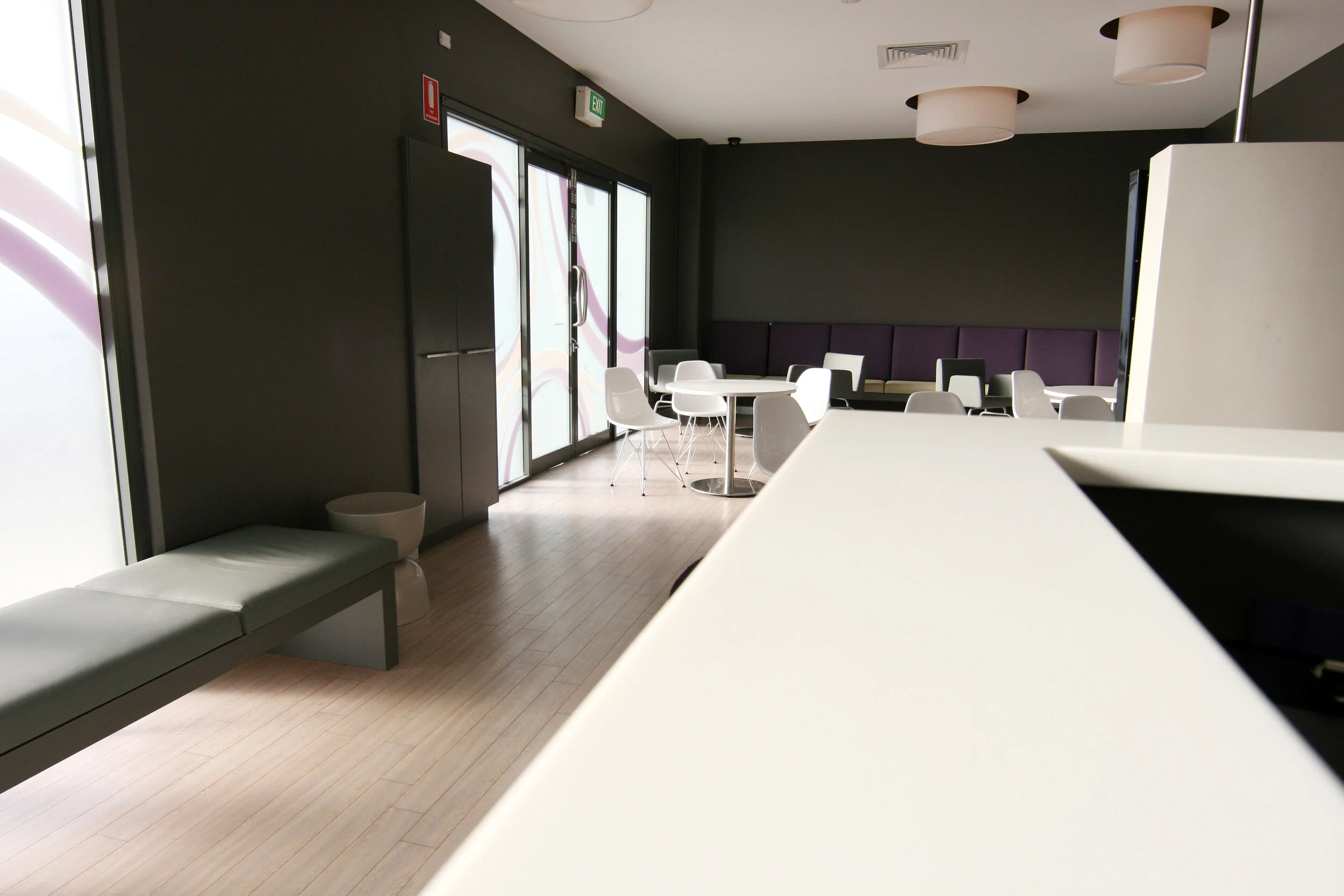 Empty modern lounge area with white chairs, round tables, dark walls, purple seating, and large windows letting in natural light.