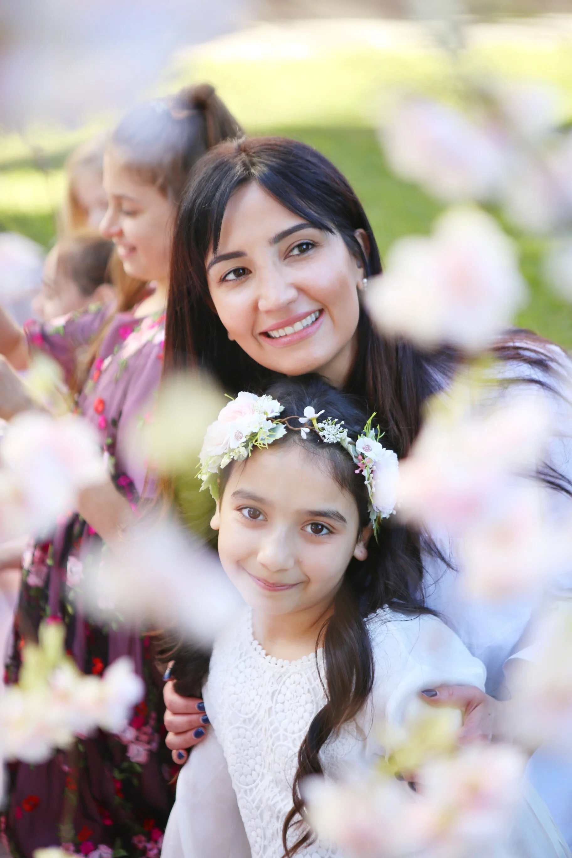 A smiling woman and a young girl with a flower crown surrounded by blurred pink and white flowers outdoors.