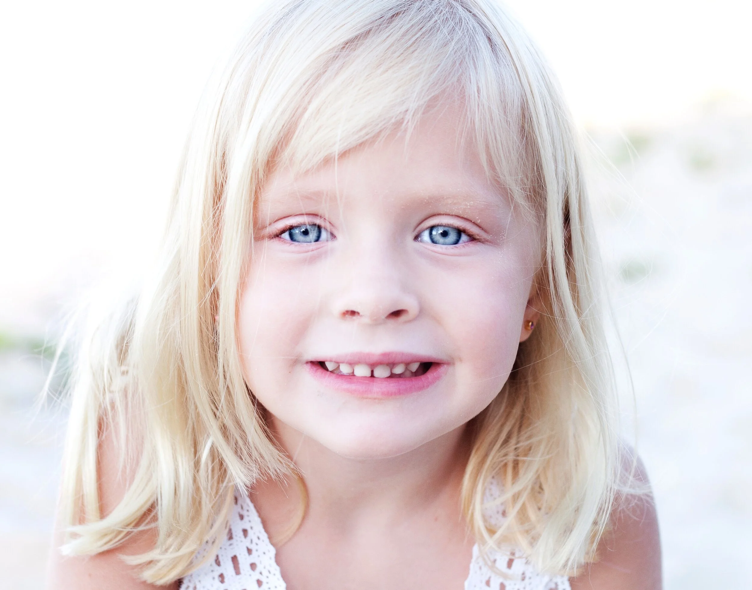 Close-up of a smiling young girl with blonde hair and blue eyes, wearing a white sleeveless top.