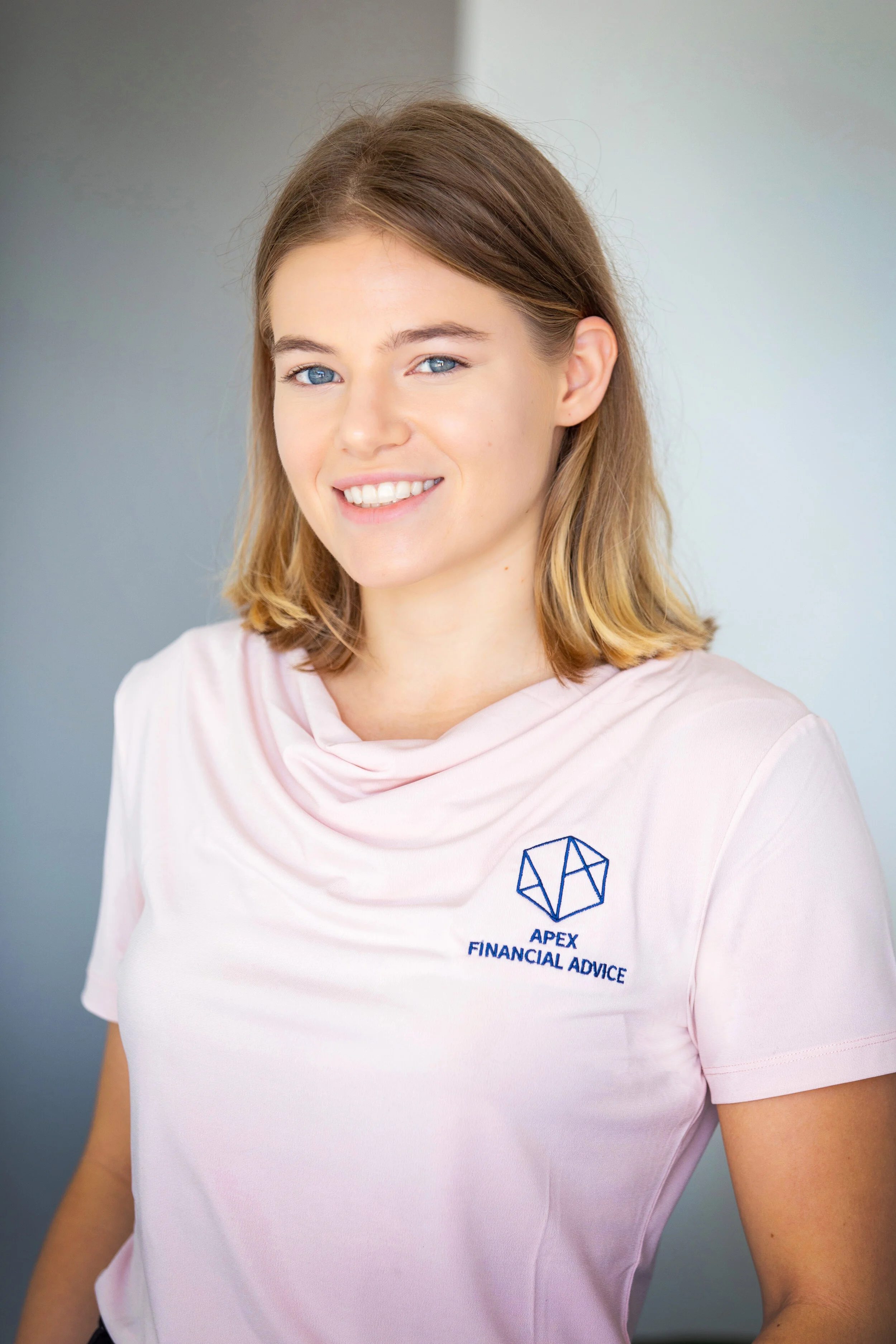 A young woman with shoulder-length blonde hair, blue eyes, and light skin smiling at the camera. She is wearing a light pink polo shirt with a logo and the text 'APEX FINANCIAL ADVICE' on it.