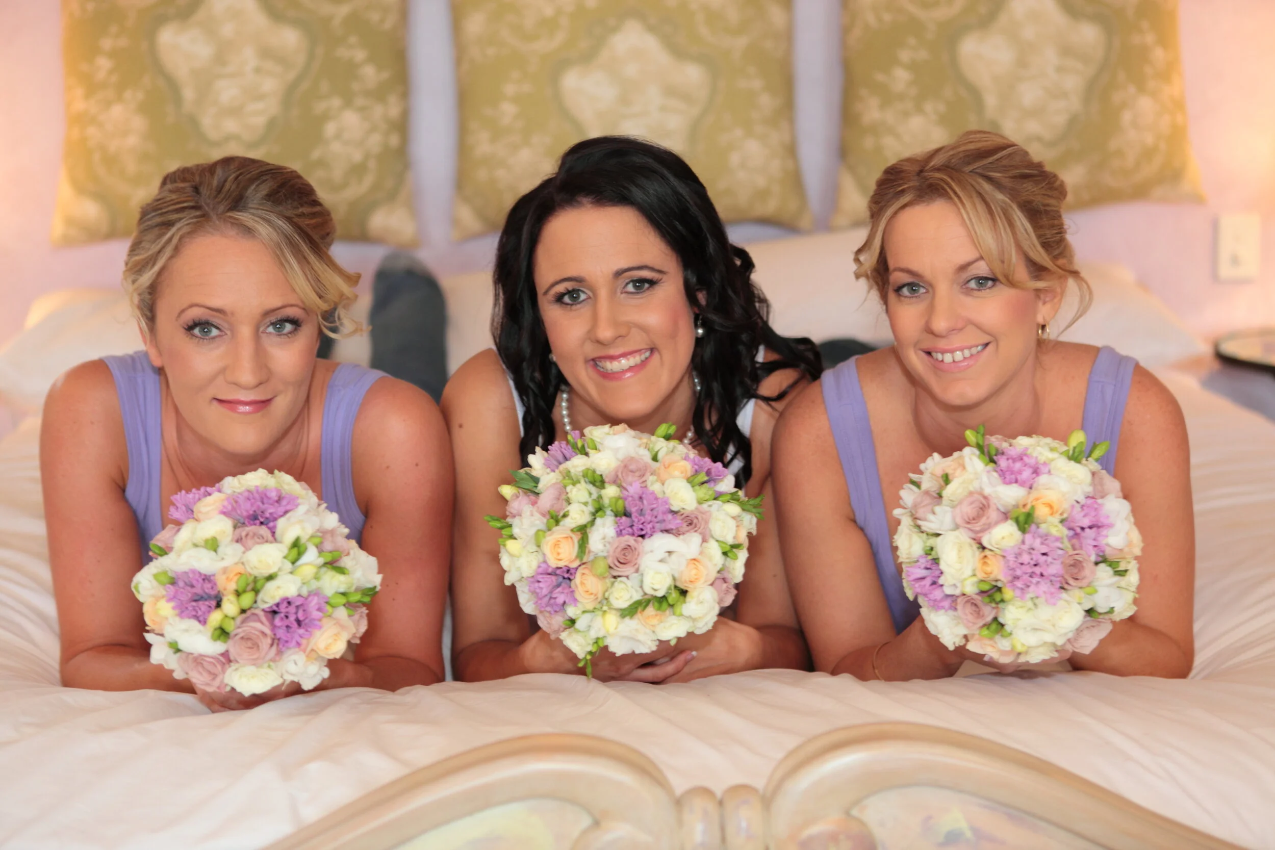 Three women lying on a bed, each holding a bouquet of pastel-colored flowers, smiling at the camera, with decorative pillows in the background.