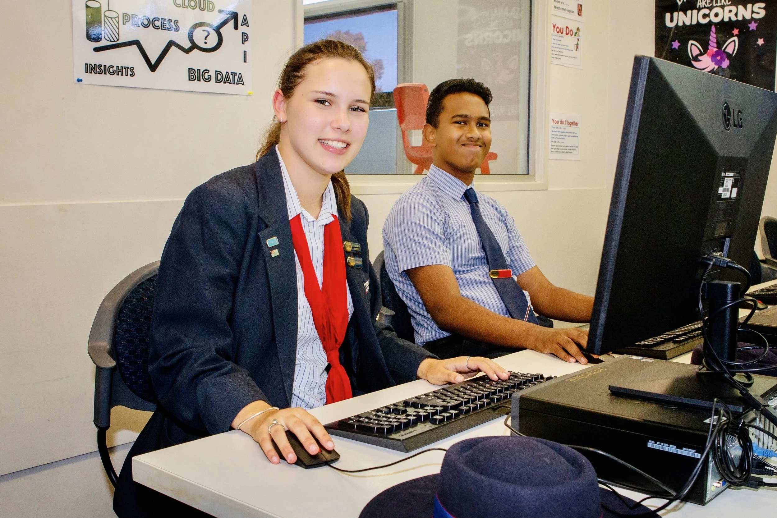 Two students, one girl with a red scarf and family blazer and one boy in a striped shirt and tie, sitting at a desk with computers in a classroom or computer lab.