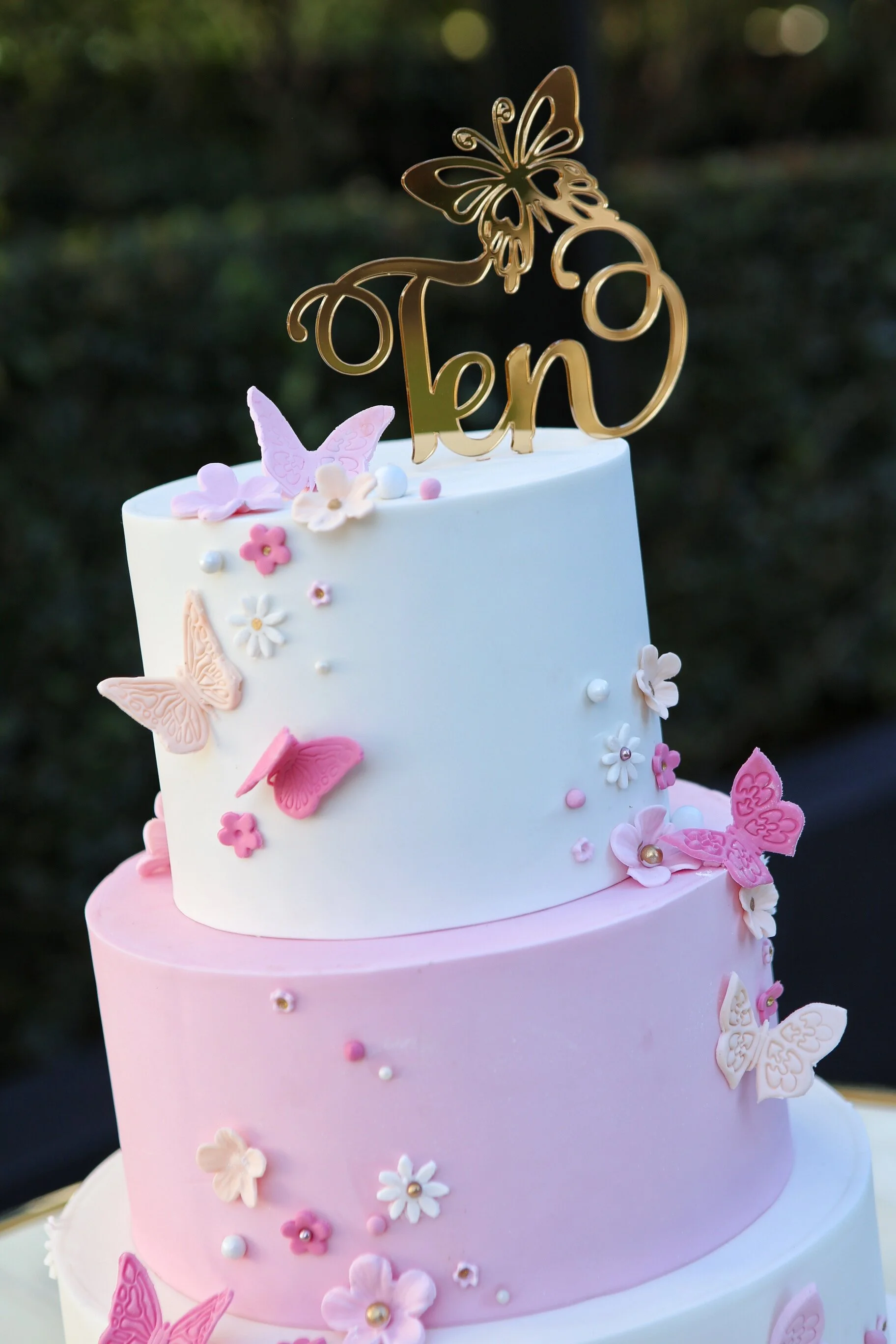 Three-tiered cake decorated with pink and white flowers and butterflies, with a gold topper that says 'Ten' with a butterfly design.