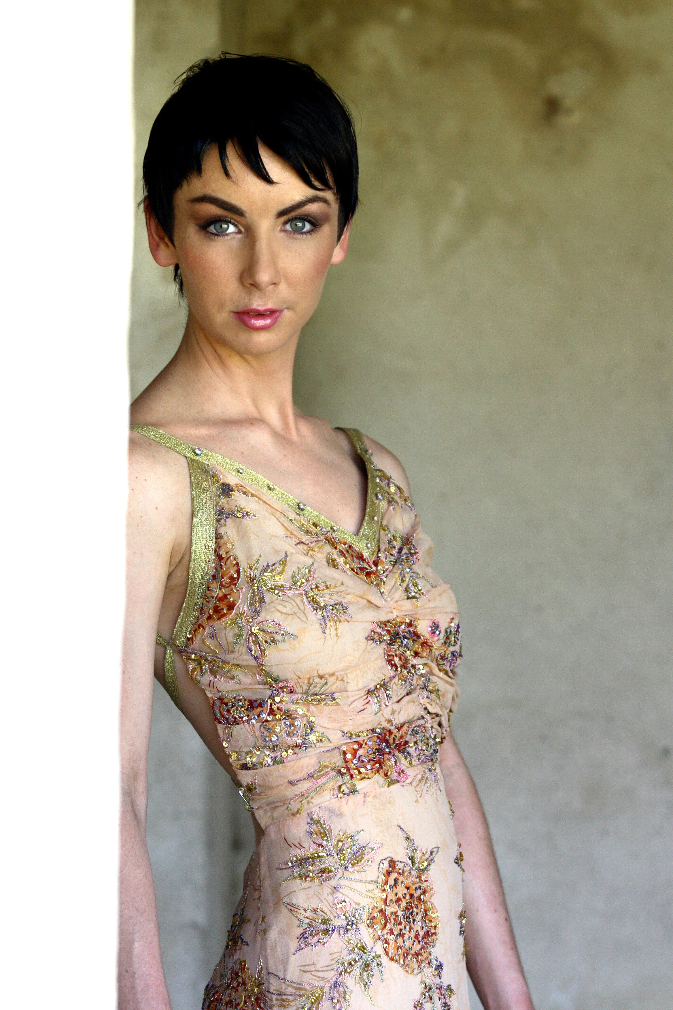 A woman with short dark hair, wearing a sleeveless, beige dress with ornate floral embroidery, standing near a wall with a neutral background.