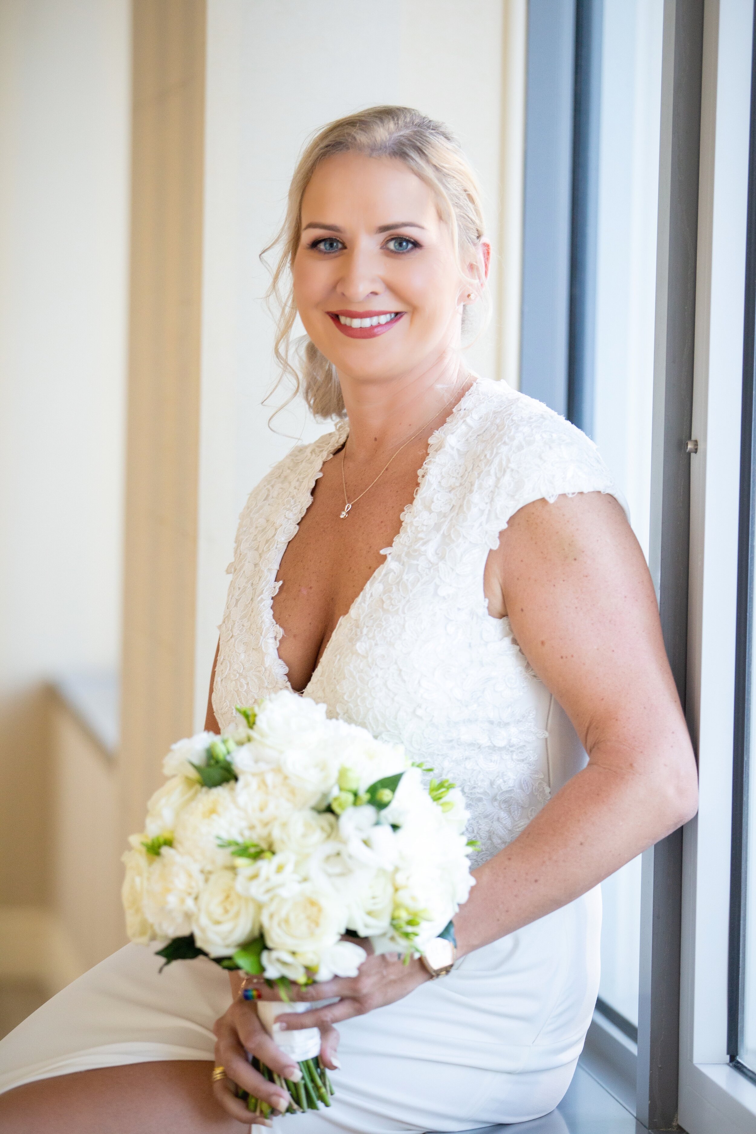 Bride in white lace dress holding a bouquet of white flowers, sitting by a window, smiling.
