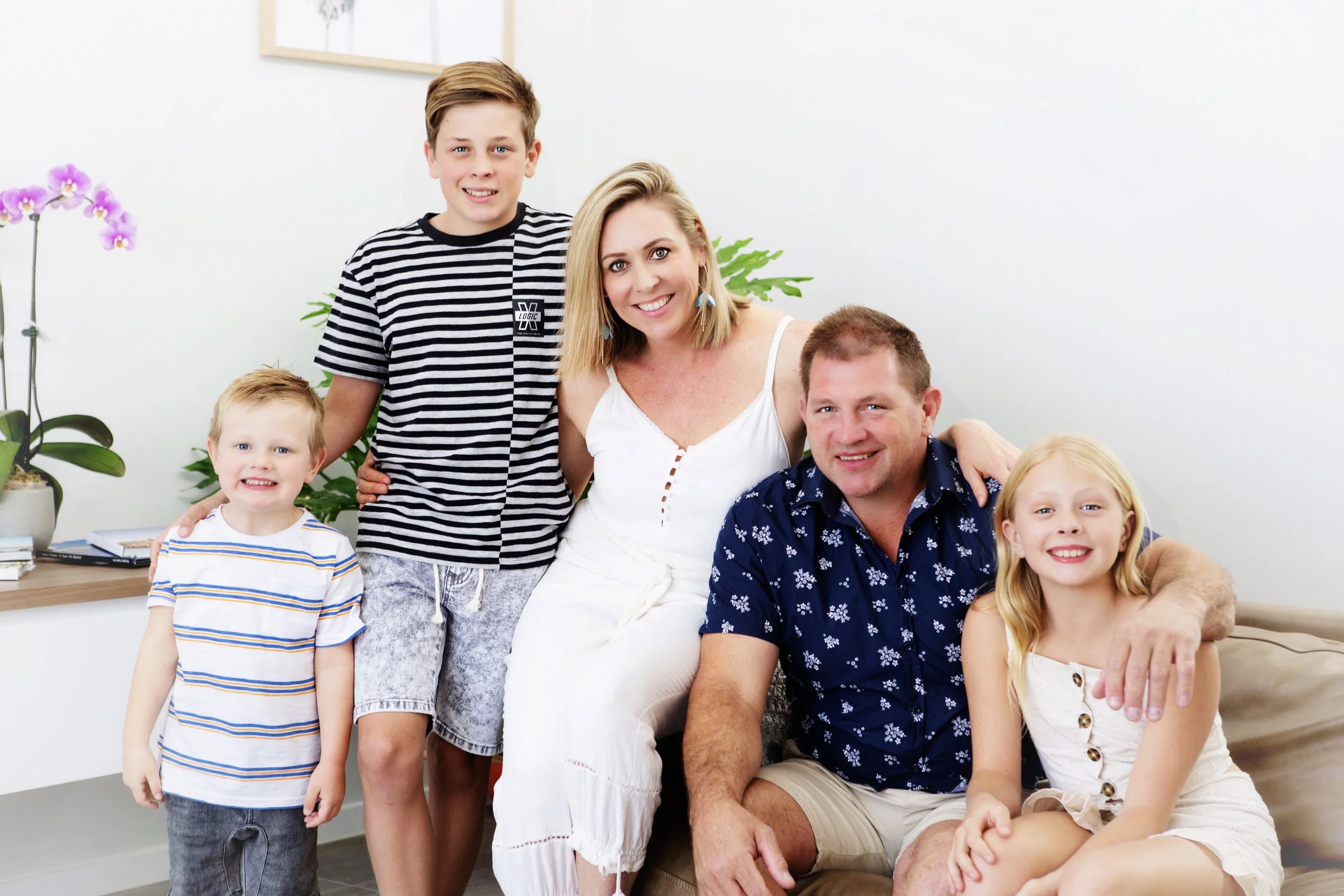 A family of six posing together indoors, smiling, with white walls, plants, and books in the background.