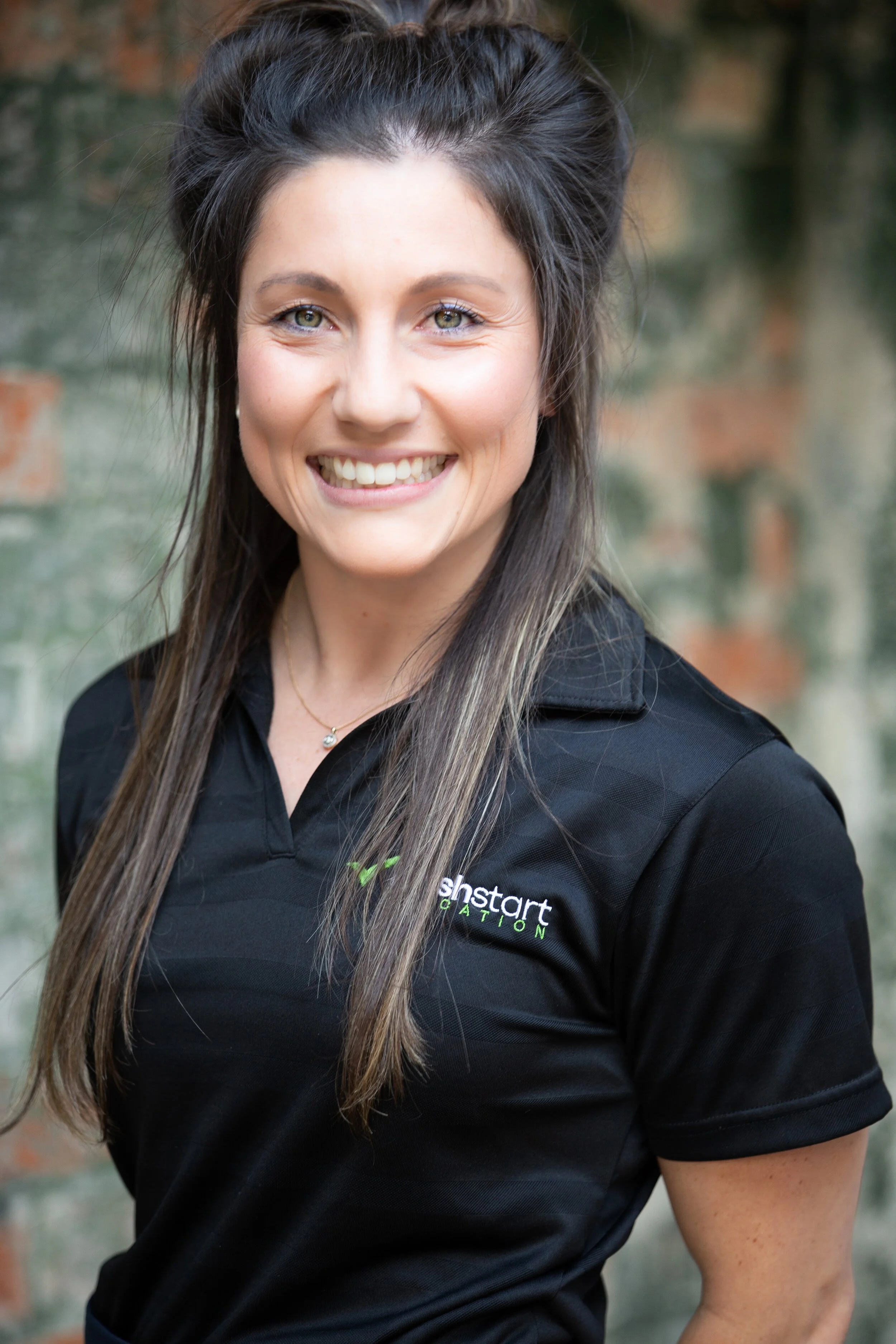 A woman smiling outdoors, wearing a black polo shirt with a logo that reads "ishstart GATION" on the chest under a leaf design, with a brick wall and greenery in the background.