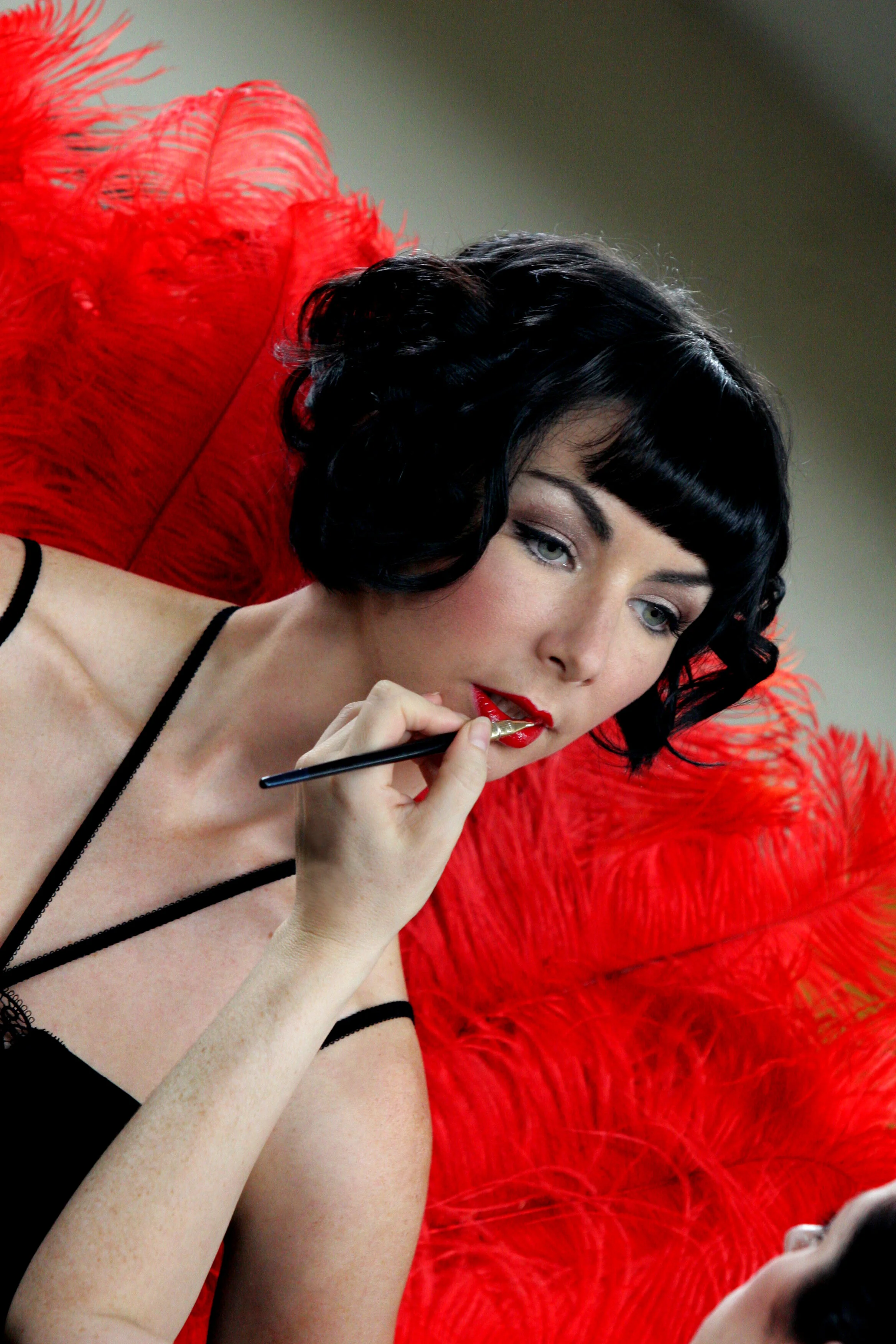 A woman with black curly hair, light skin, and red lipstick shades her lips with a small brush while sitting in front of a vibrant red feathered backdrop.