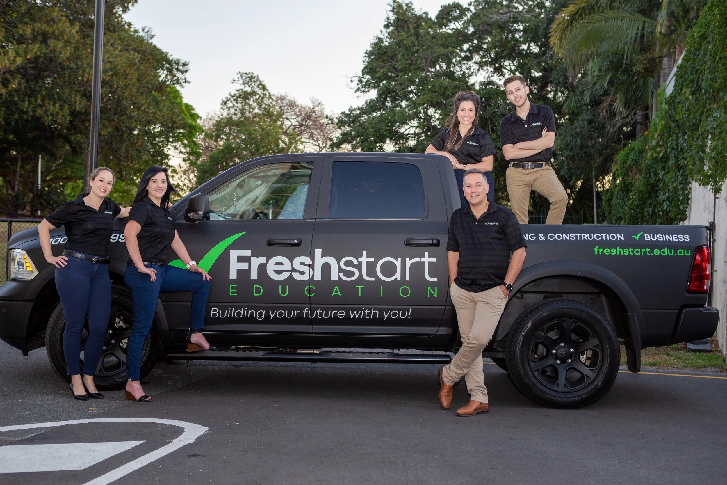 Group of six people, four women and two men, posing around a black pickup truck with 'Freshstart Education' branding, parked outdoors with trees in the background.