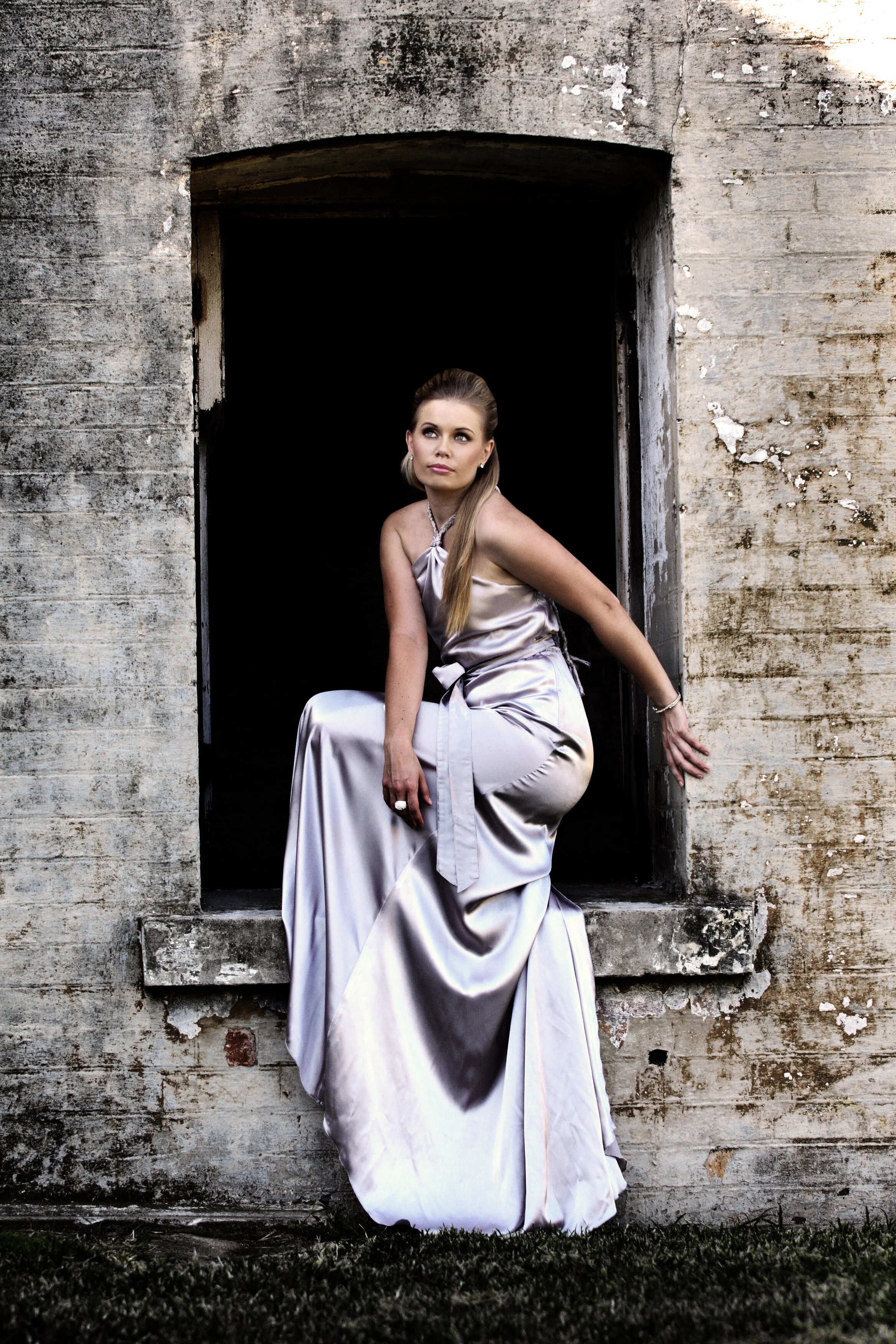 A woman in a silver satin dress sitting on a windowsill outside a weathered brick building, looking off to the side.
