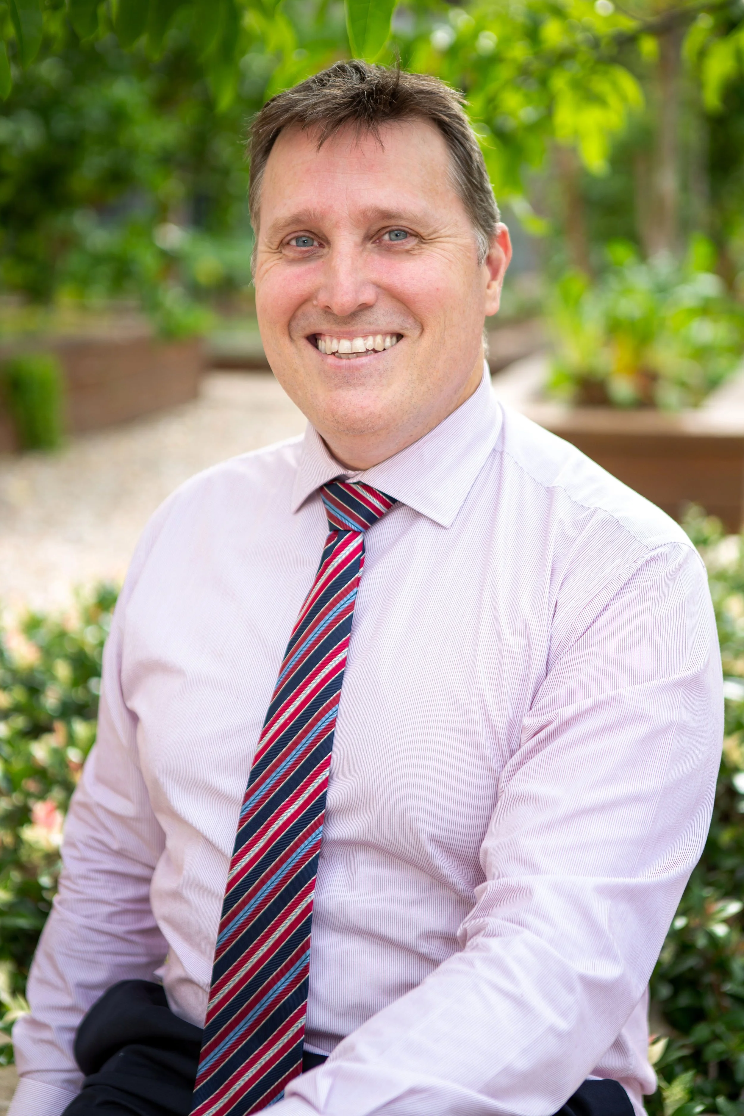 Portrait of a smiling man in a light pink shirt and striped tie, sitting outdoors with green foliage in the background.