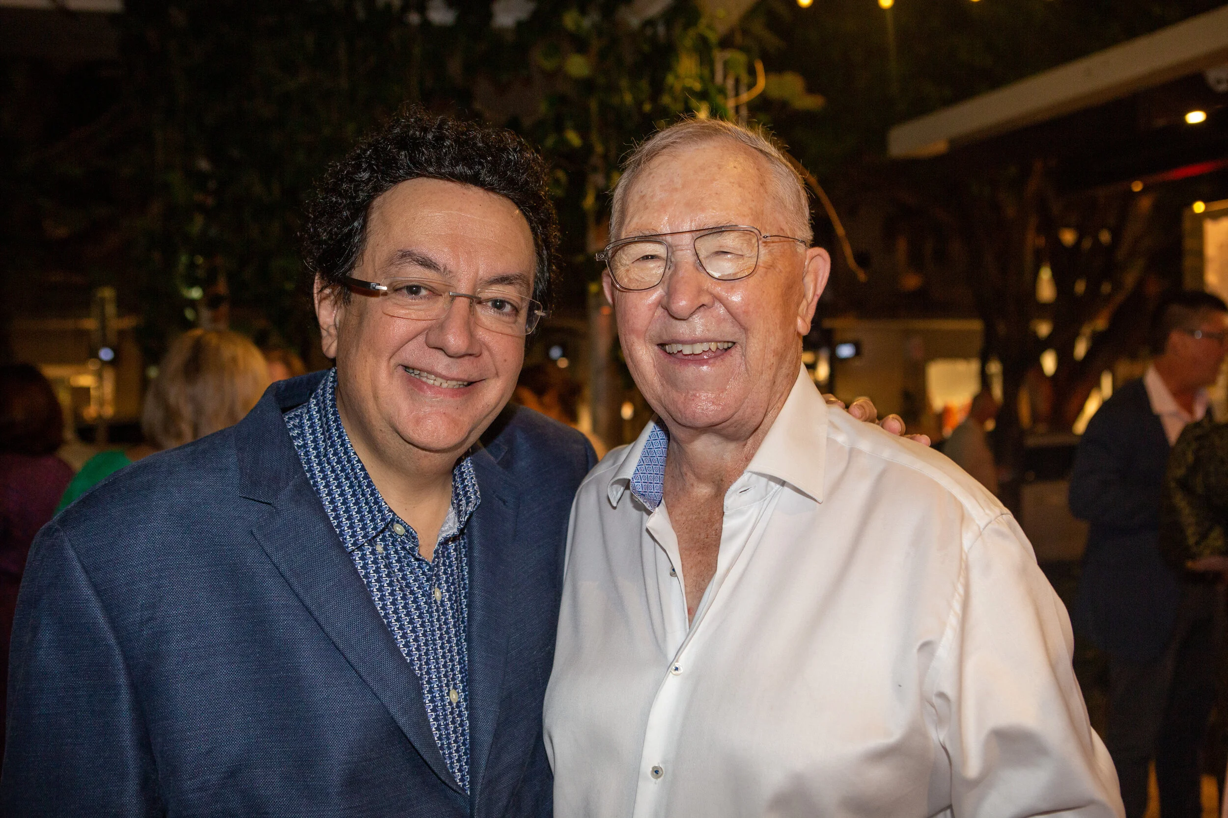 Two smiling men in formal attire at a social event, standing close together with arms around each other, outdoors at night with trees and other people in the background.