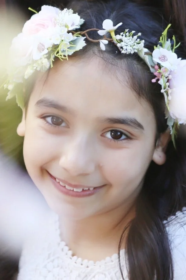 Close-up of a young girl with a floral crown, smiling, with dark hair and big brown eyes.