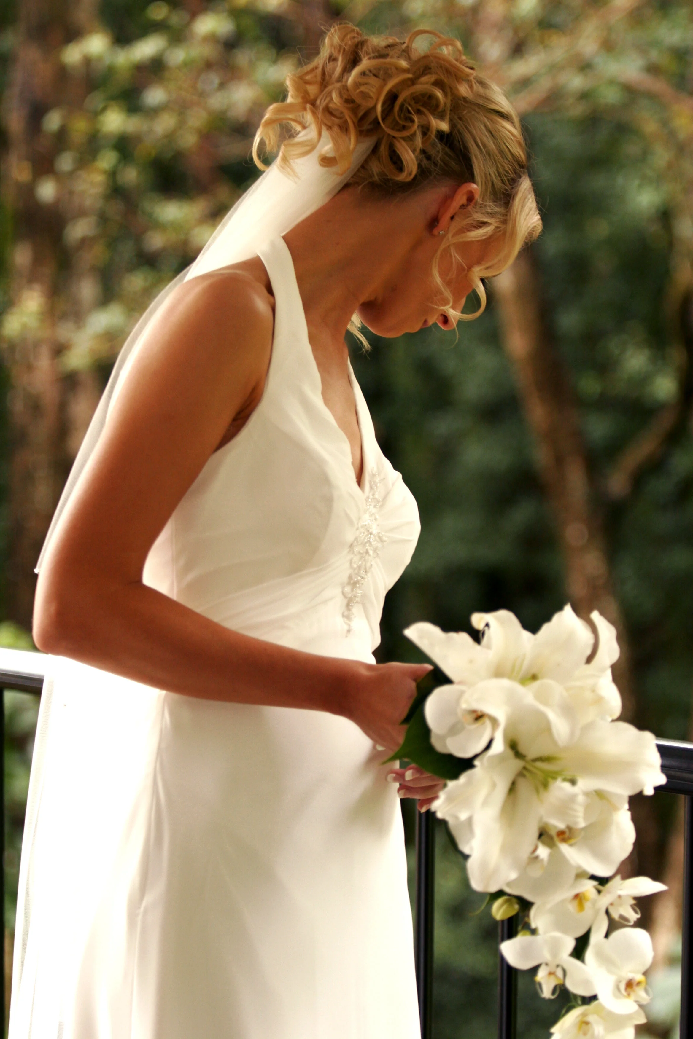 Bride in a white wedding dress holding a bouquet of white lilies, standing outdoors amid trees.