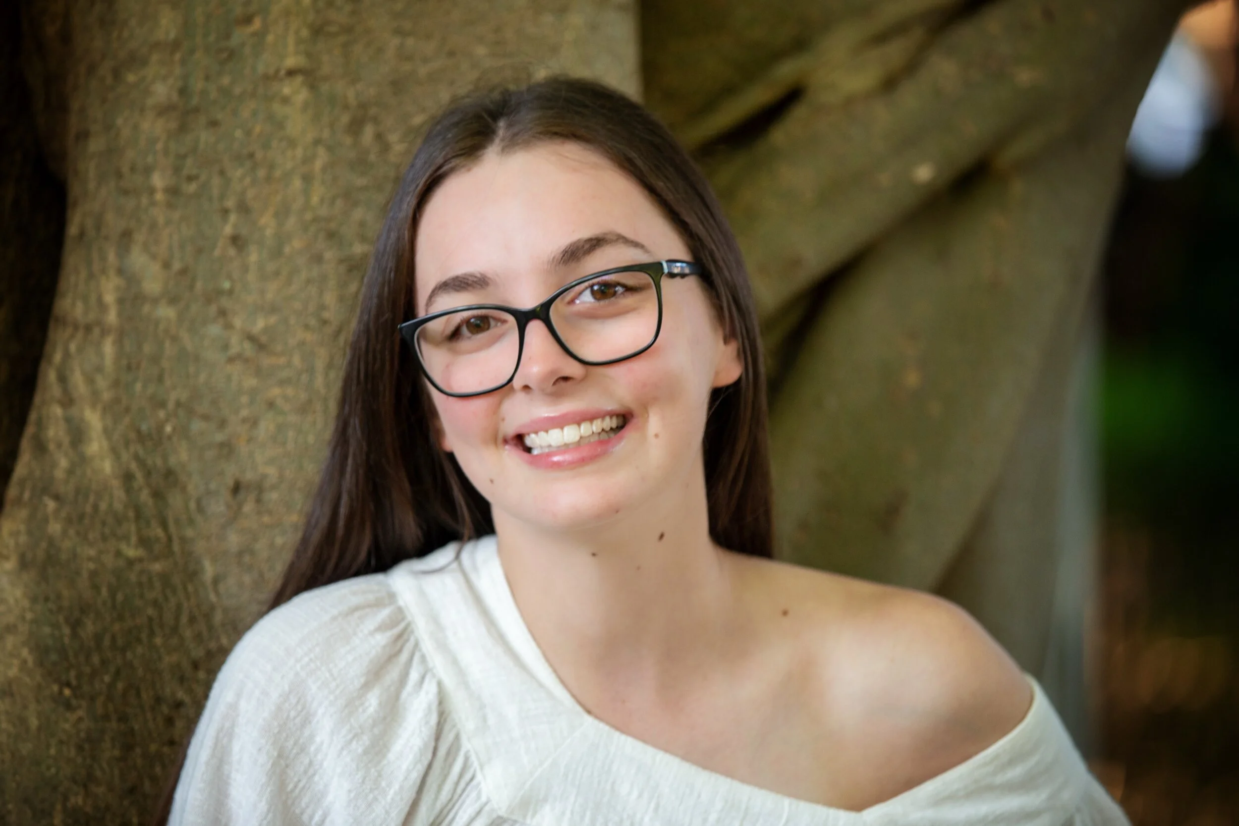 A young woman with long brown hair, glasses, and a white off-shoulder top smiling outdoors in front of a tree.