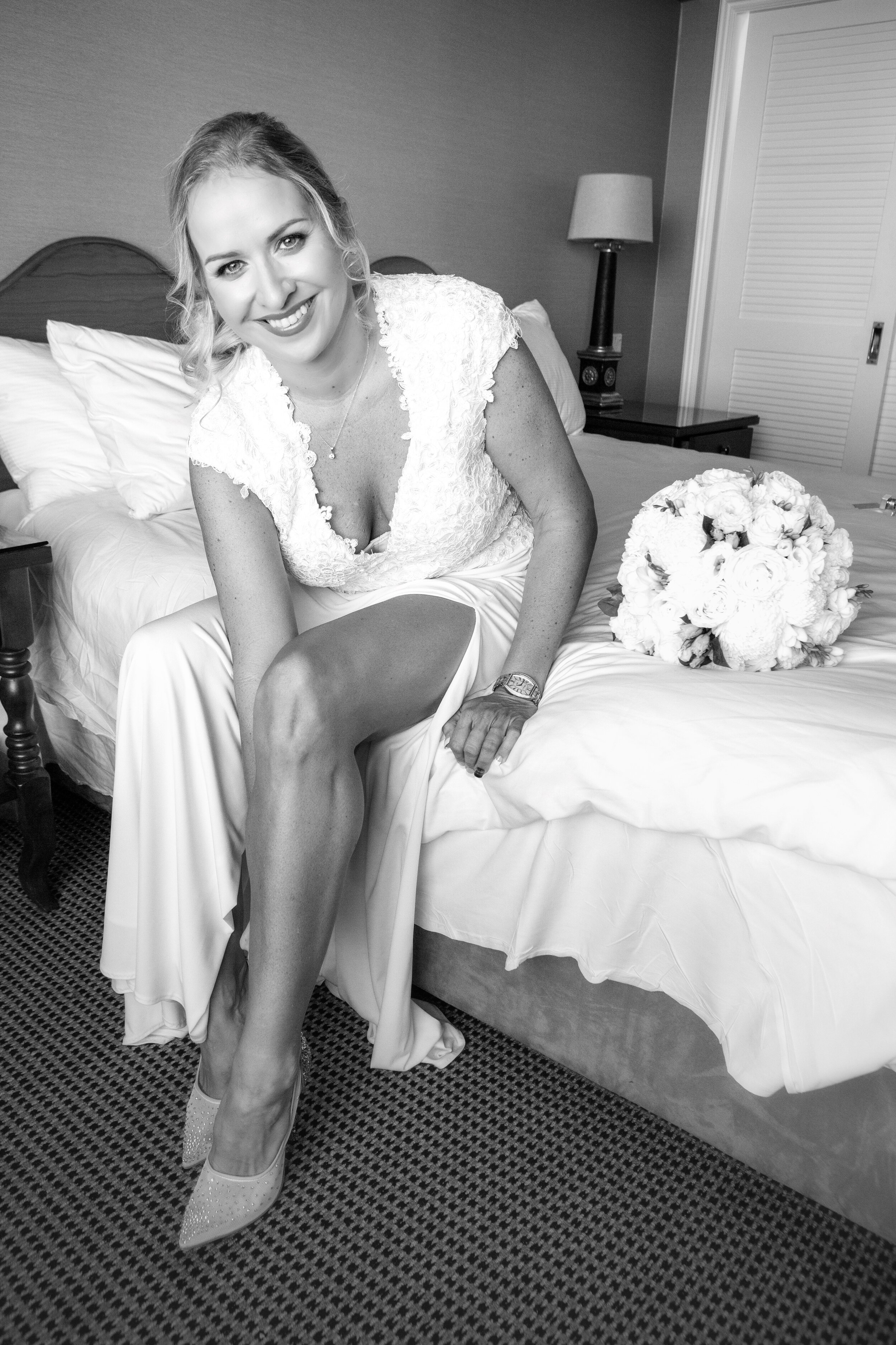 A woman in a wedding dress sitting on a bed, smiling, with a bouquet of flowers nearby.