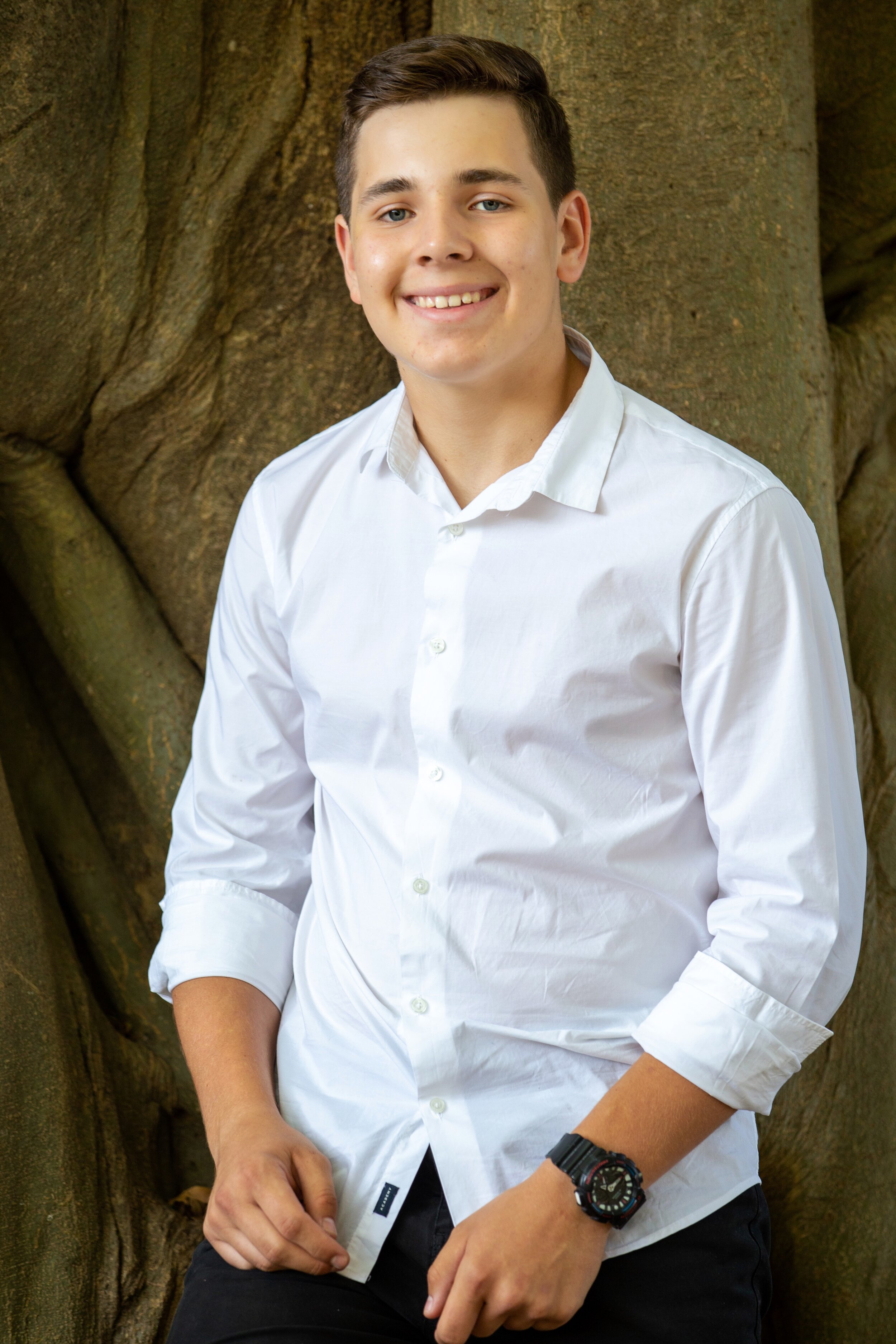 A young man wearing a white button-up shirt and black pants, standing outdoors in front of a large tree, smiling at the camera.