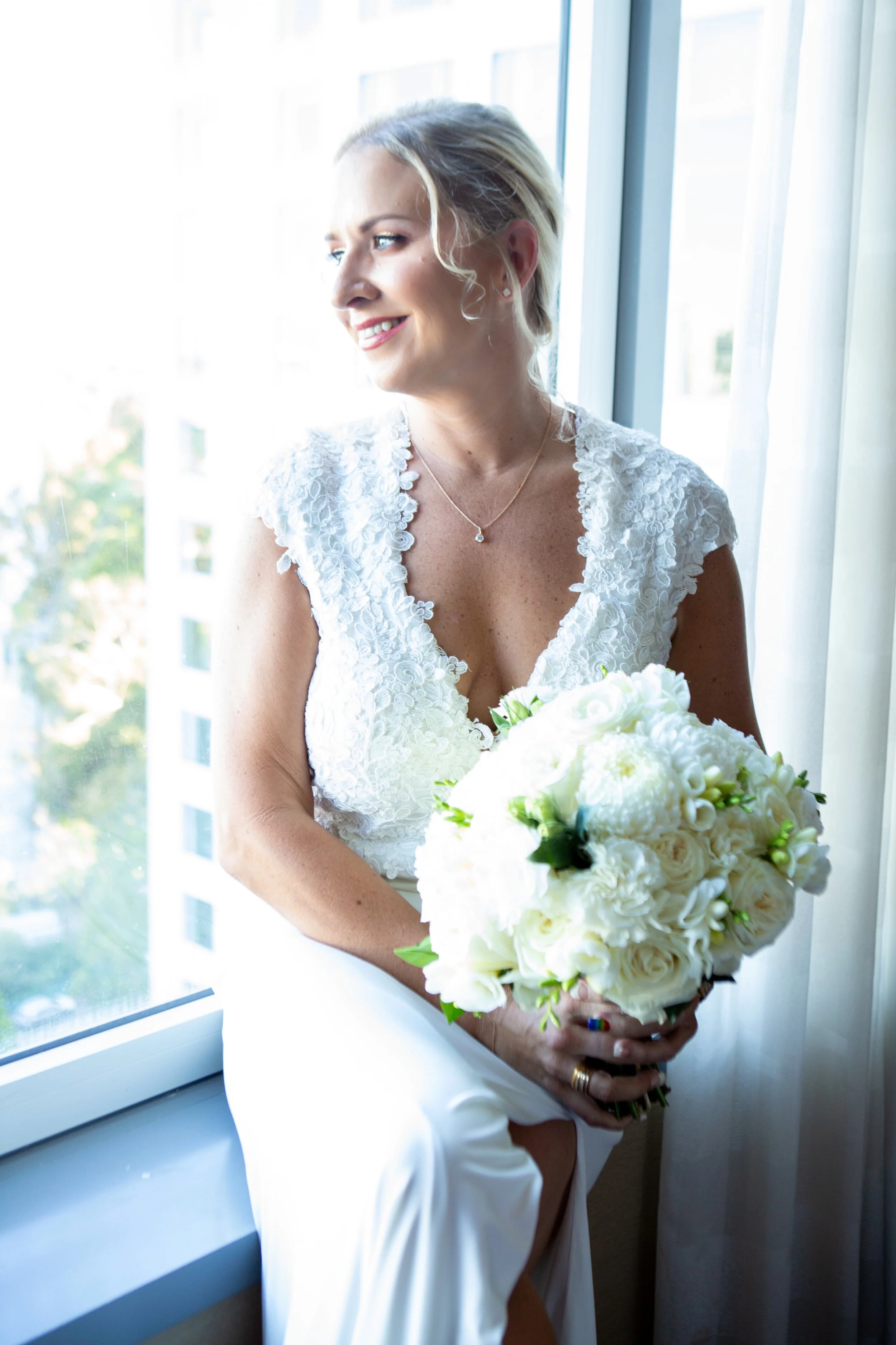 A smiling bride dressed in white holding a bouquet of white flowers, sitting by a window with natural light.