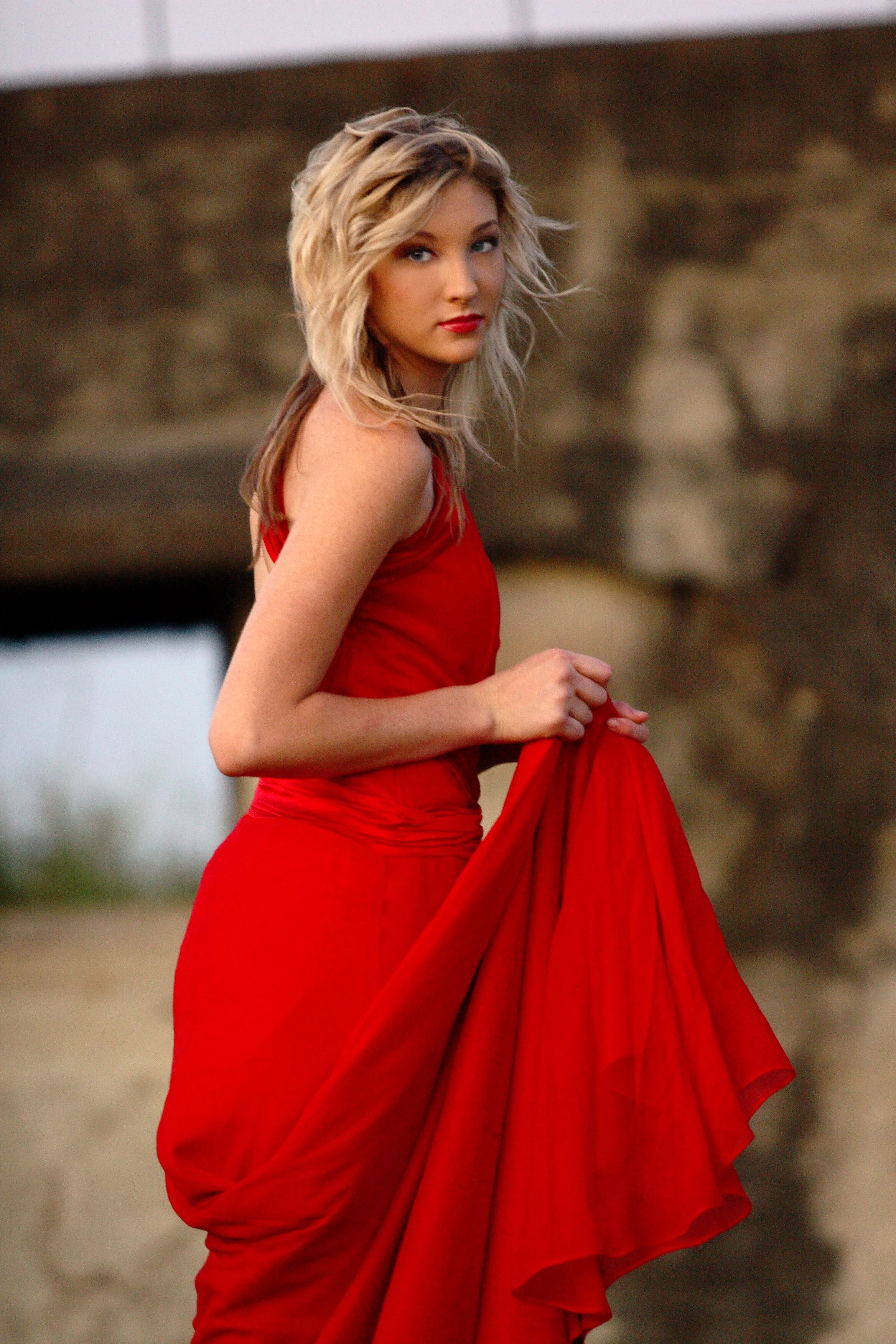 A young woman with blonde wavy hair, wearing a red dress, holding the skirt with her right hand, standing outdoors near a stone bridge or wall.