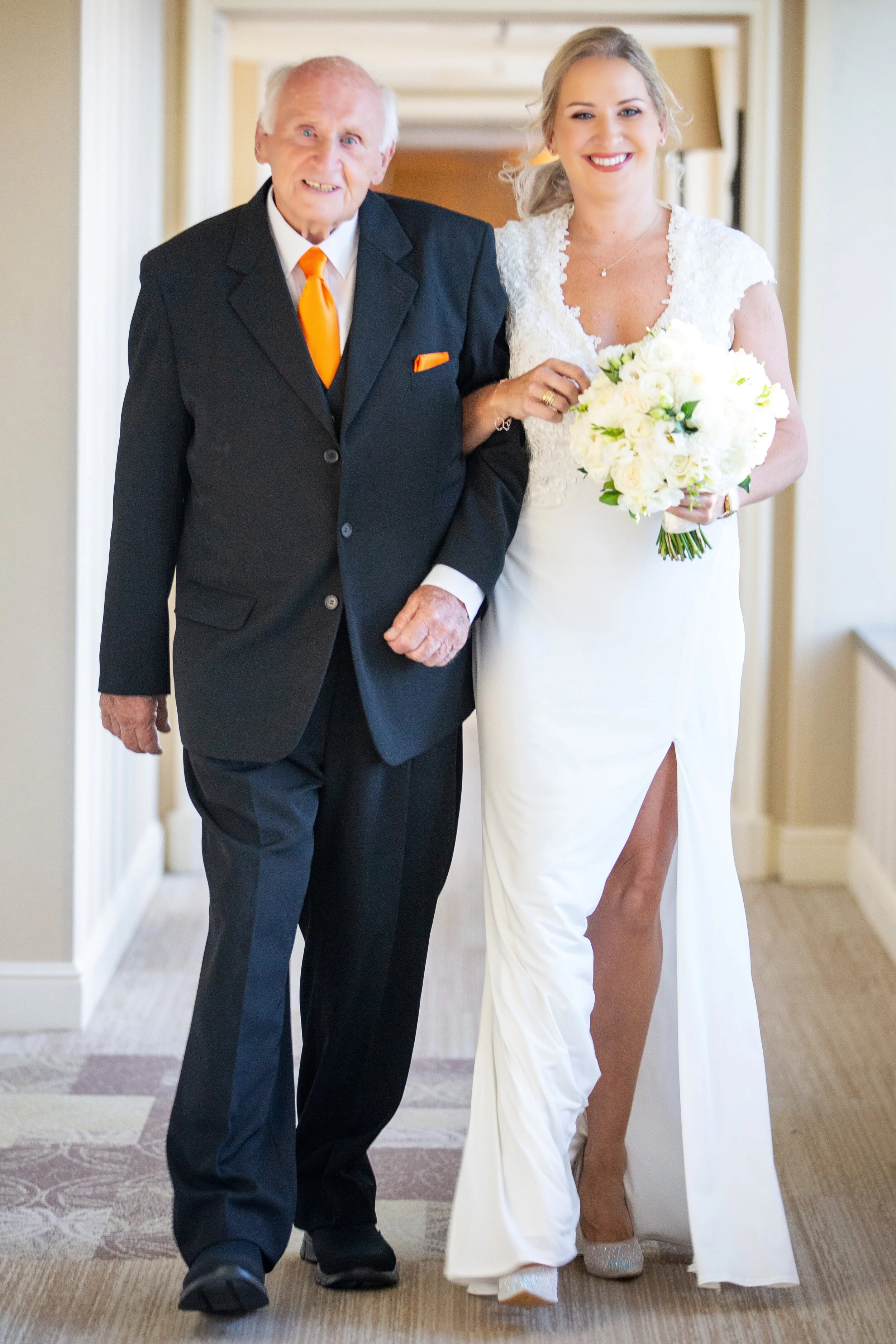 An elderly man in a black suit with an orange tie walking arm-in-arm with a smiling woman in a white wedding dress holding a bouquet of white flowers.