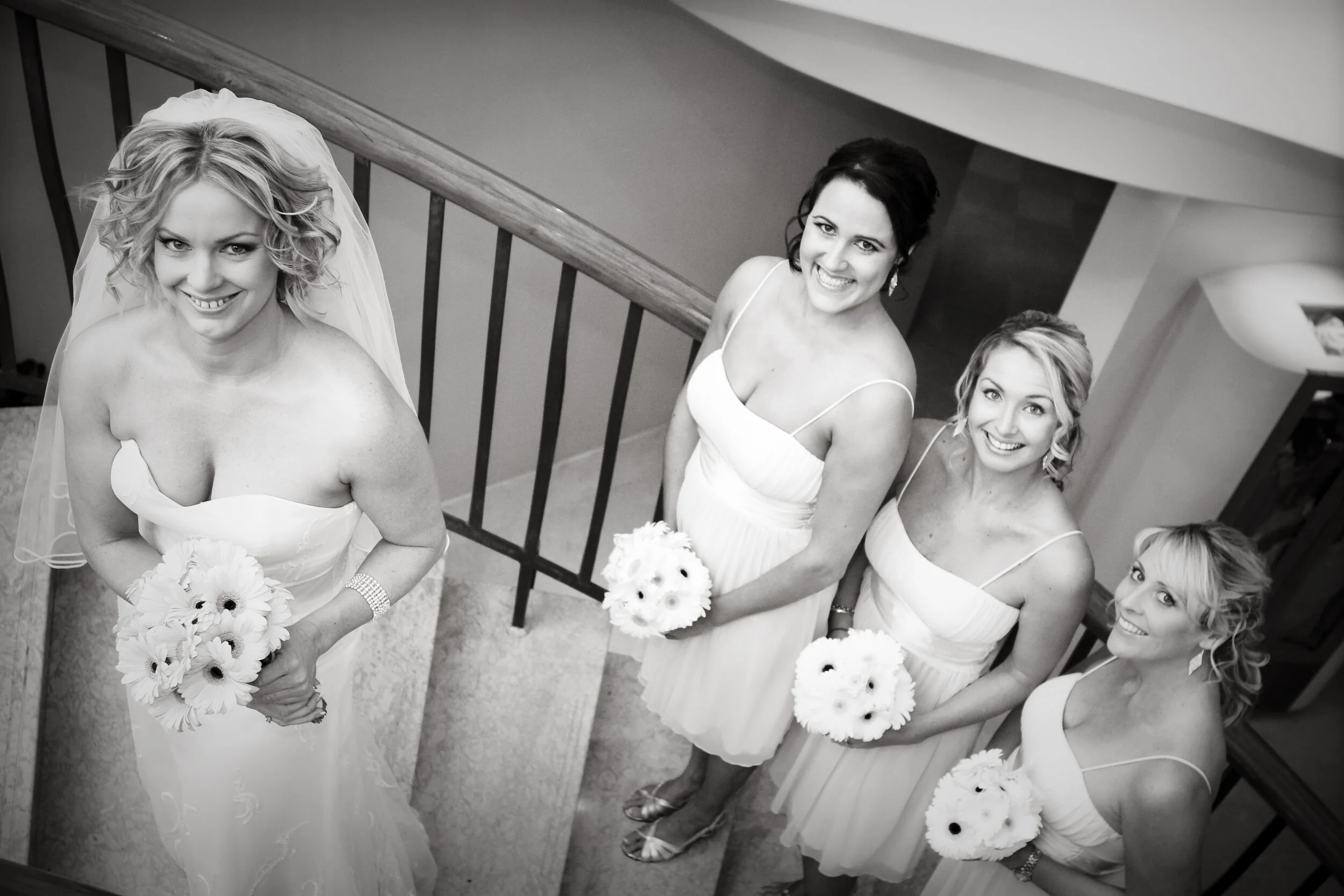 Black and white photo of four women dressed in white, smiling and holding bouquets, standing on a staircase, with one woman wearing a wedding dress and the others in bridesmaid dresses.